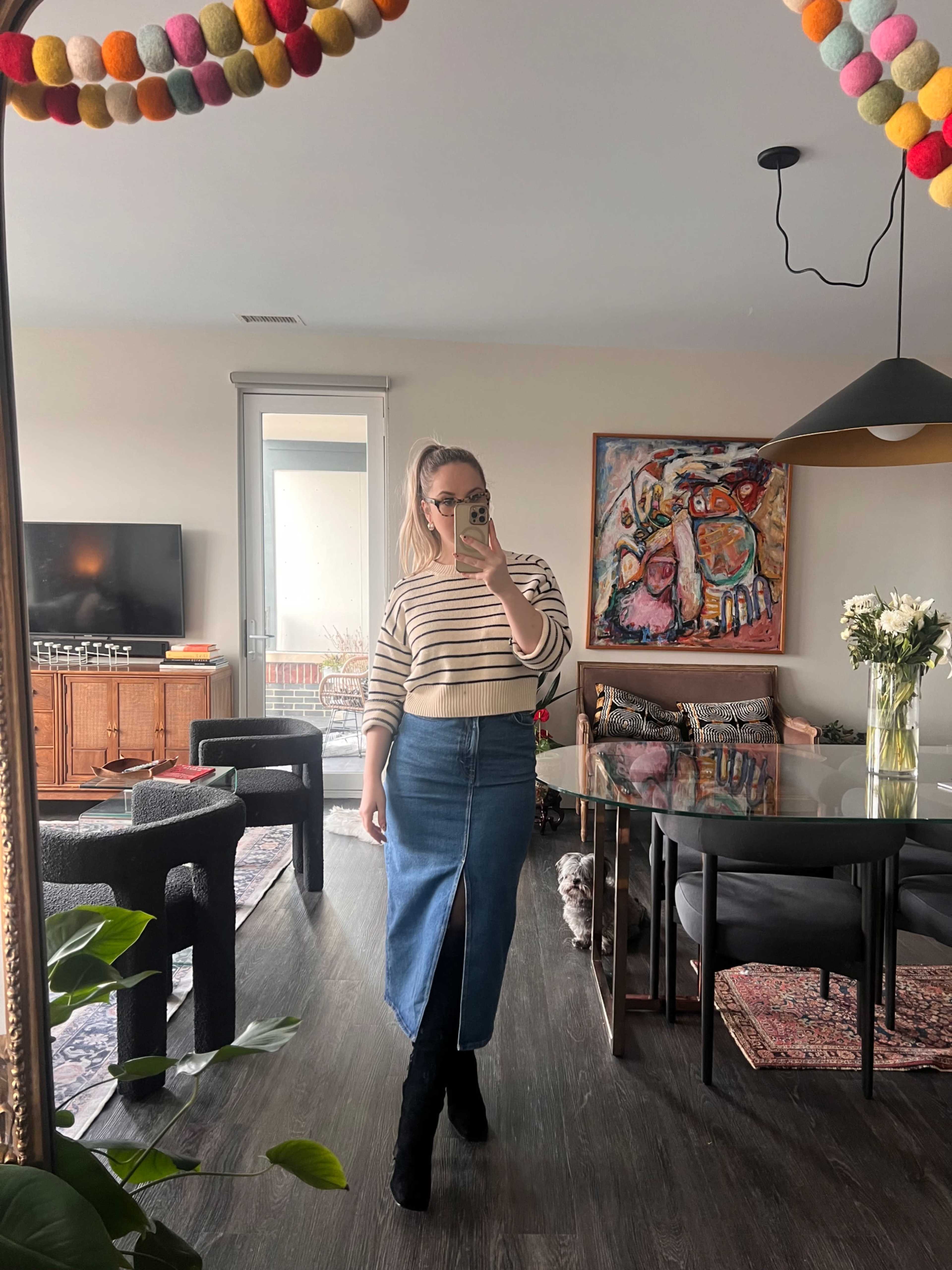 A person stands in a well-decorated living room, wearing a striped sweater and a denim skirt, with a mirror reflecting the space that includes a television and a dining table.