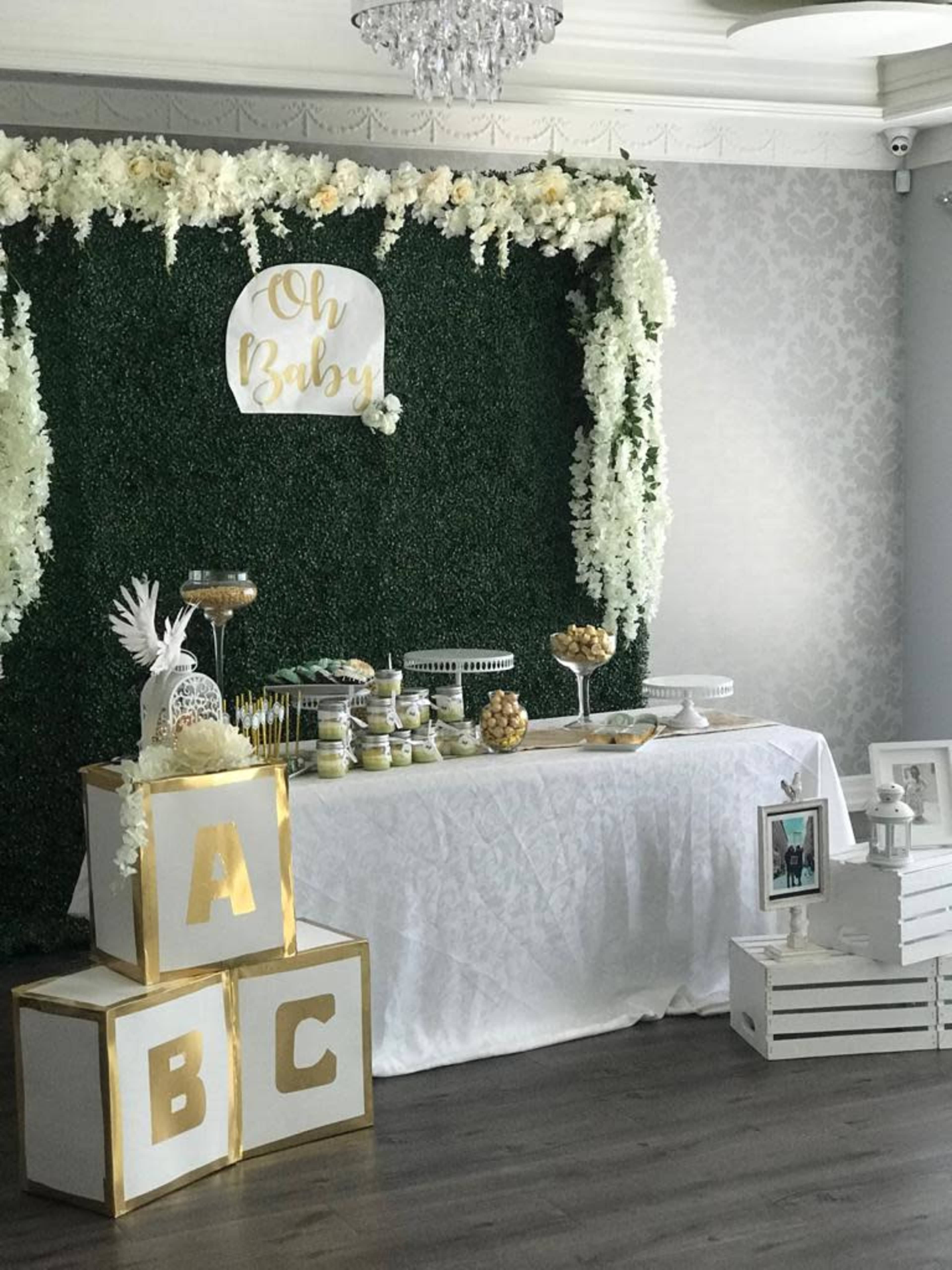 A decorated event space with a green floral backdrop, a table covered with treats, and decorative blocks labeled "A," "B," and "C" in front of the table.