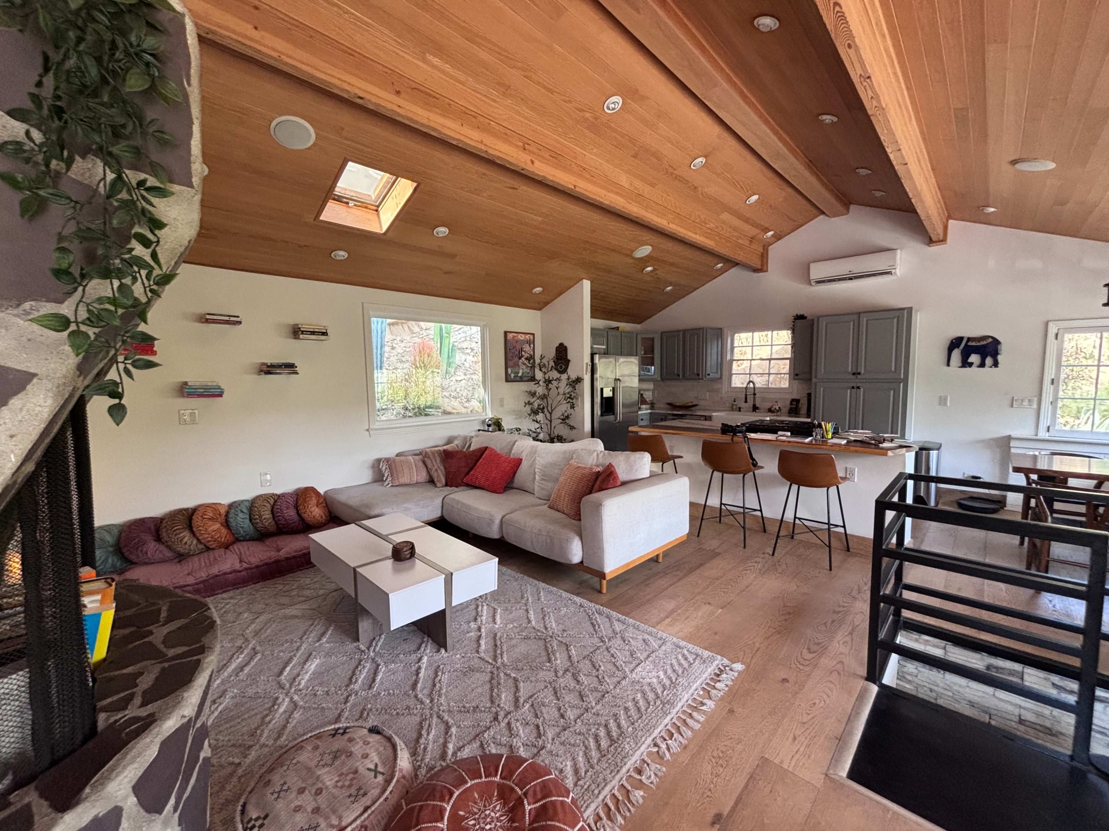 The image shows a modern living space with a sectional sofa, a coffee table, and an open kitchen area, featuring wooden beams and large windows.