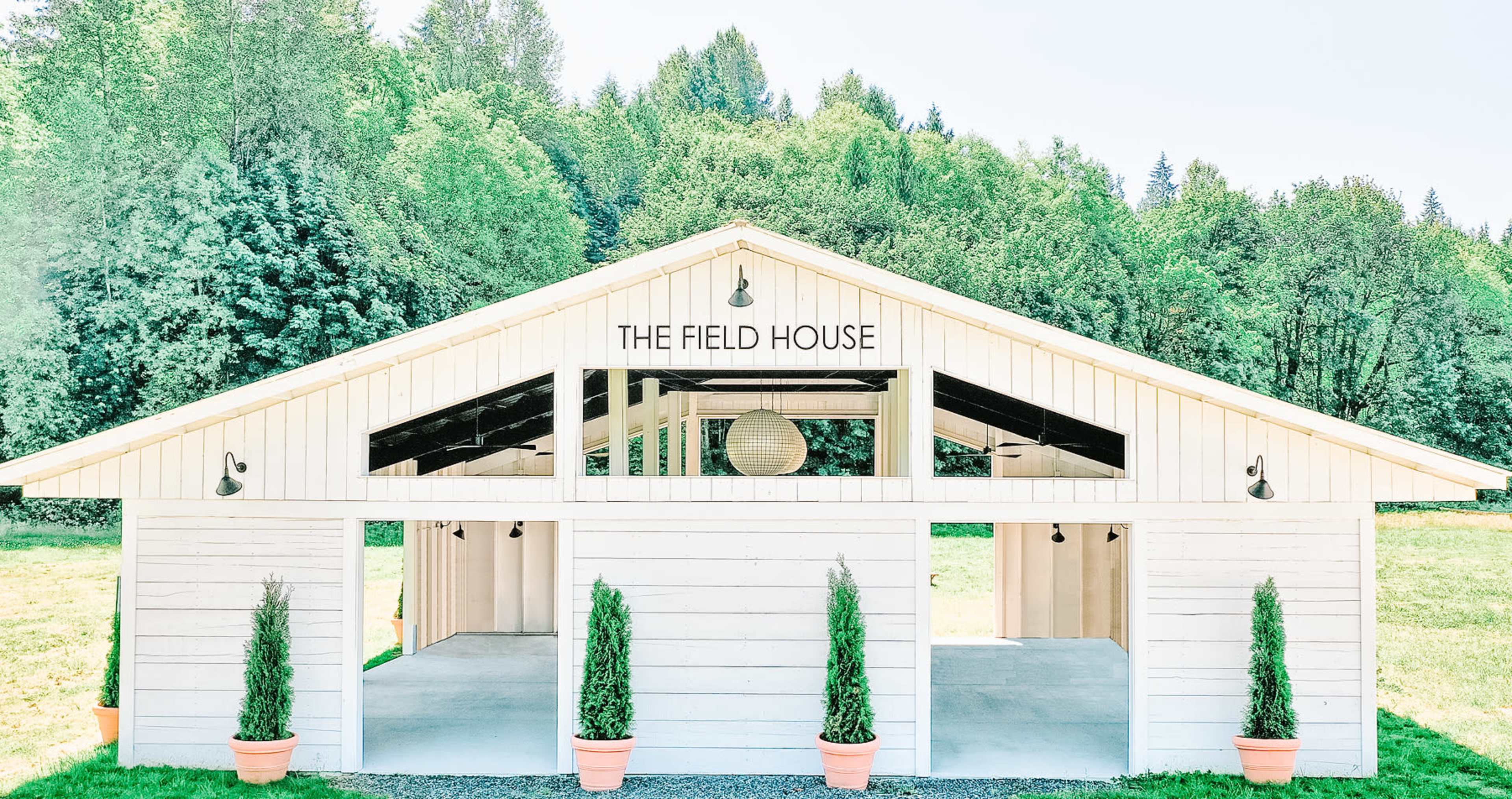 A large, white wooden building named "The Field House" with three open sections at the front and potted plants on either side, set against a backdrop of greenery.