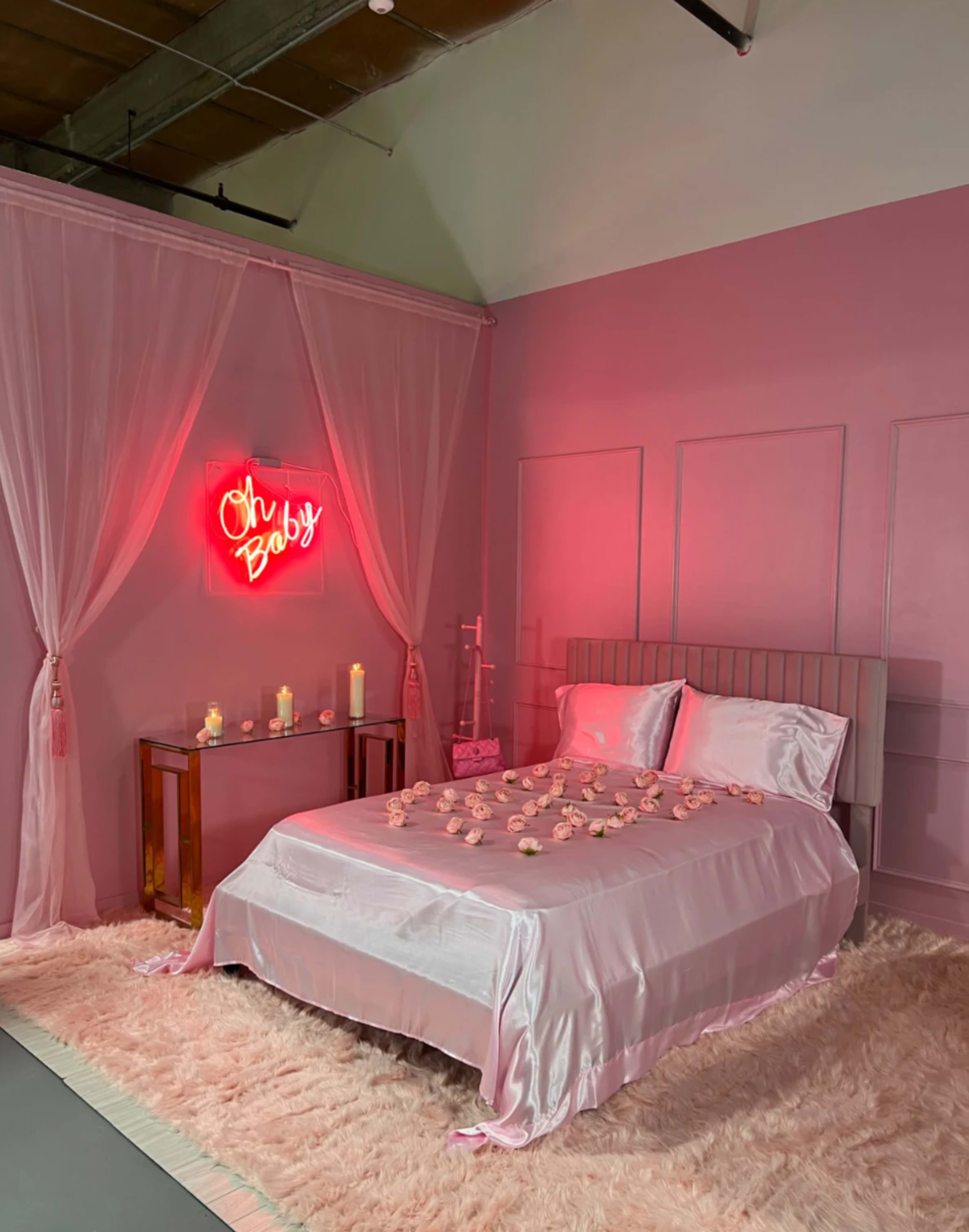 A pink-themed bedroom features a bed covered in satin sheets, with rose petals scattered on top, adorned by a neon sign that reads "Oh Baby" and surrounded by candles.