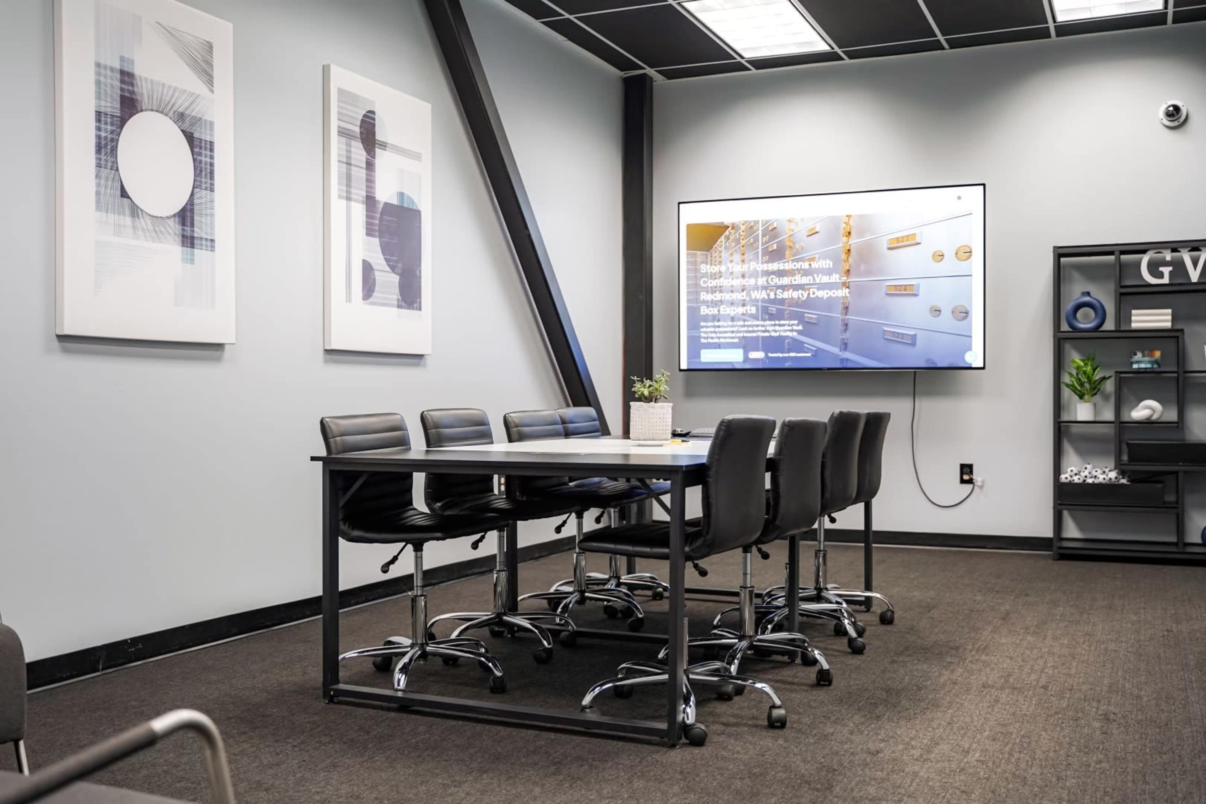 A modern conference room features a long table surrounded by black chairs and a large screen displaying a presentation on the wall.