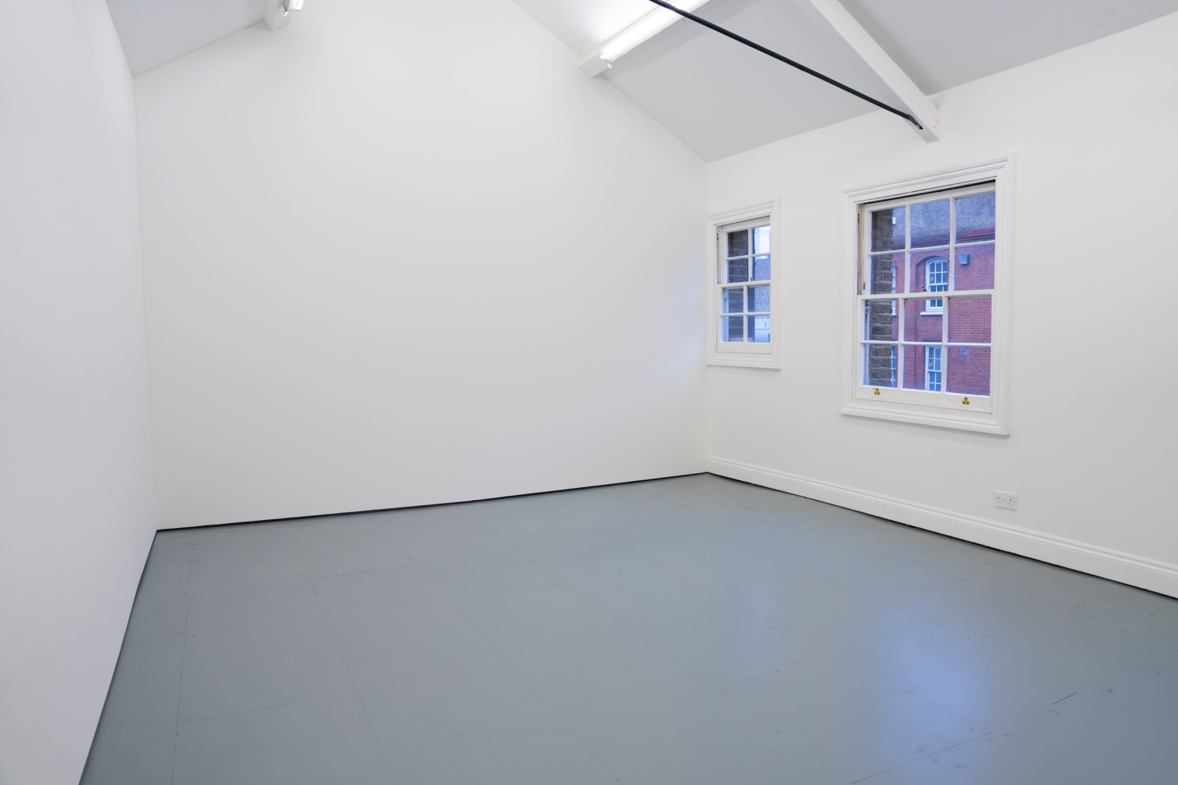 The image shows a minimalistic, empty room with white walls, a gray floor, and two windows facing an exterior brick wall.