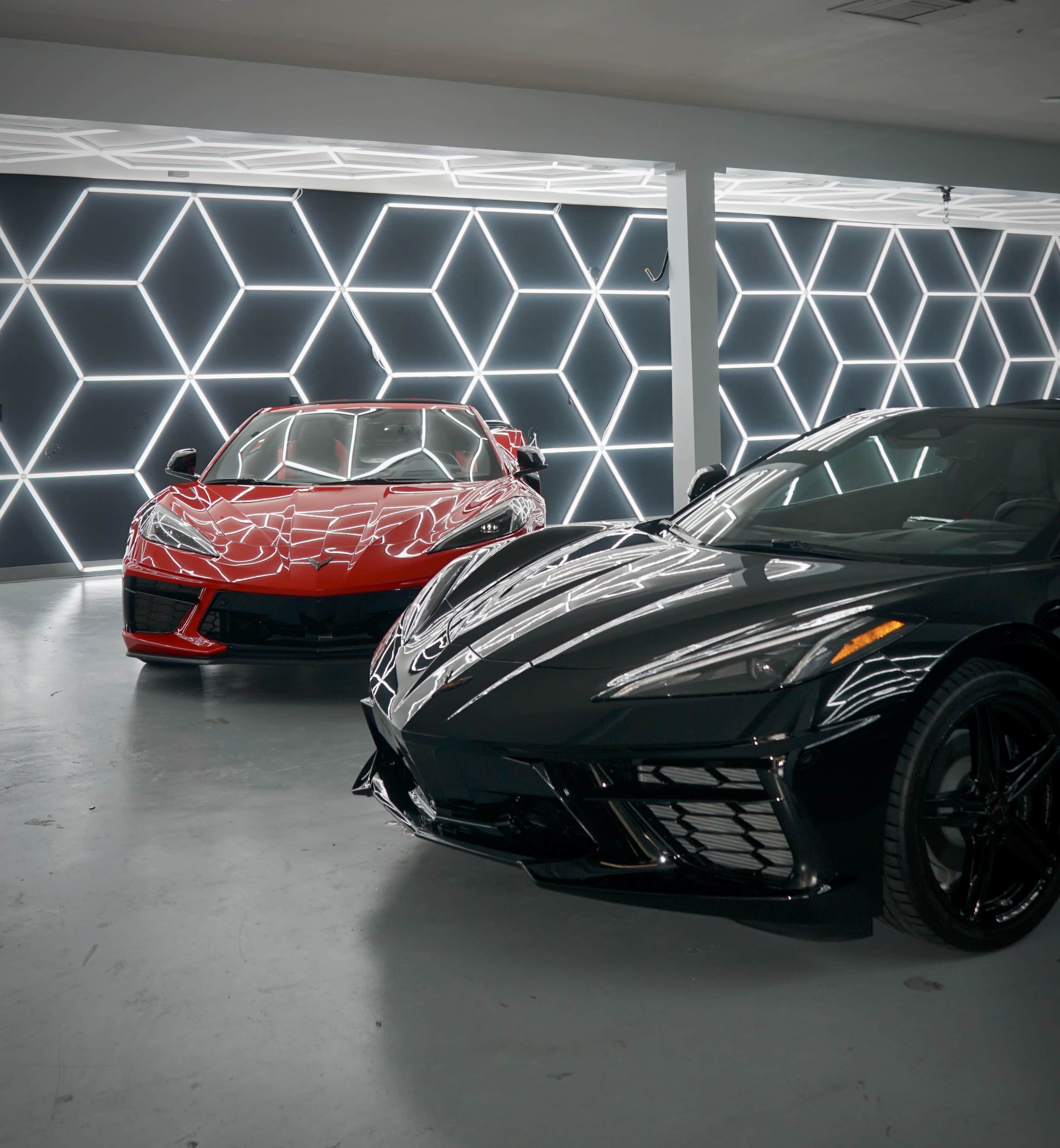 Two sleek sports cars, one red and one black, are parked in a modern showroom with a geometric light installation on the wall behind them.