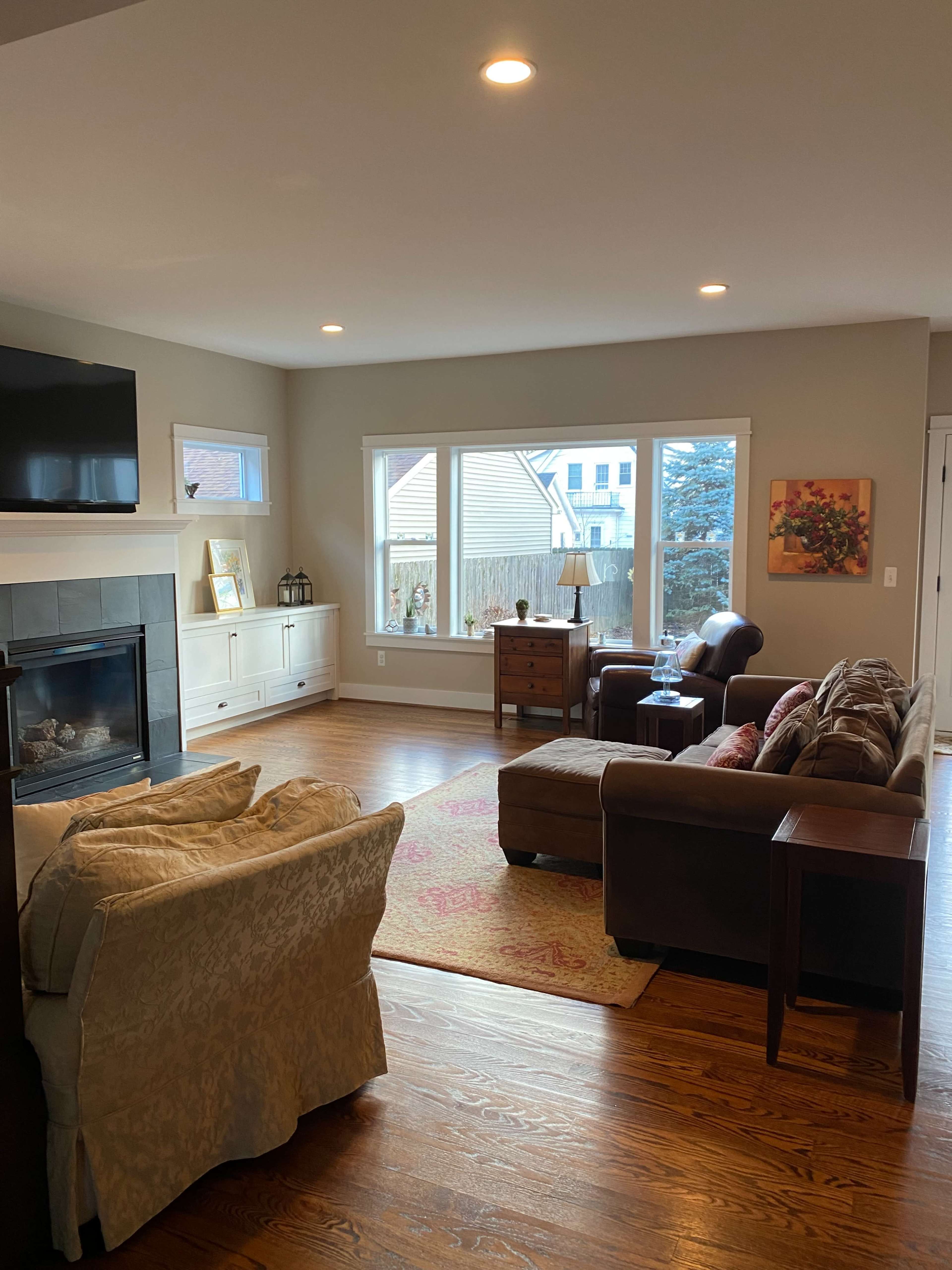 The image shows a living room with a fireplace, a sofa, an armchair, a wooden coffee table, and large windows allowing natural light.