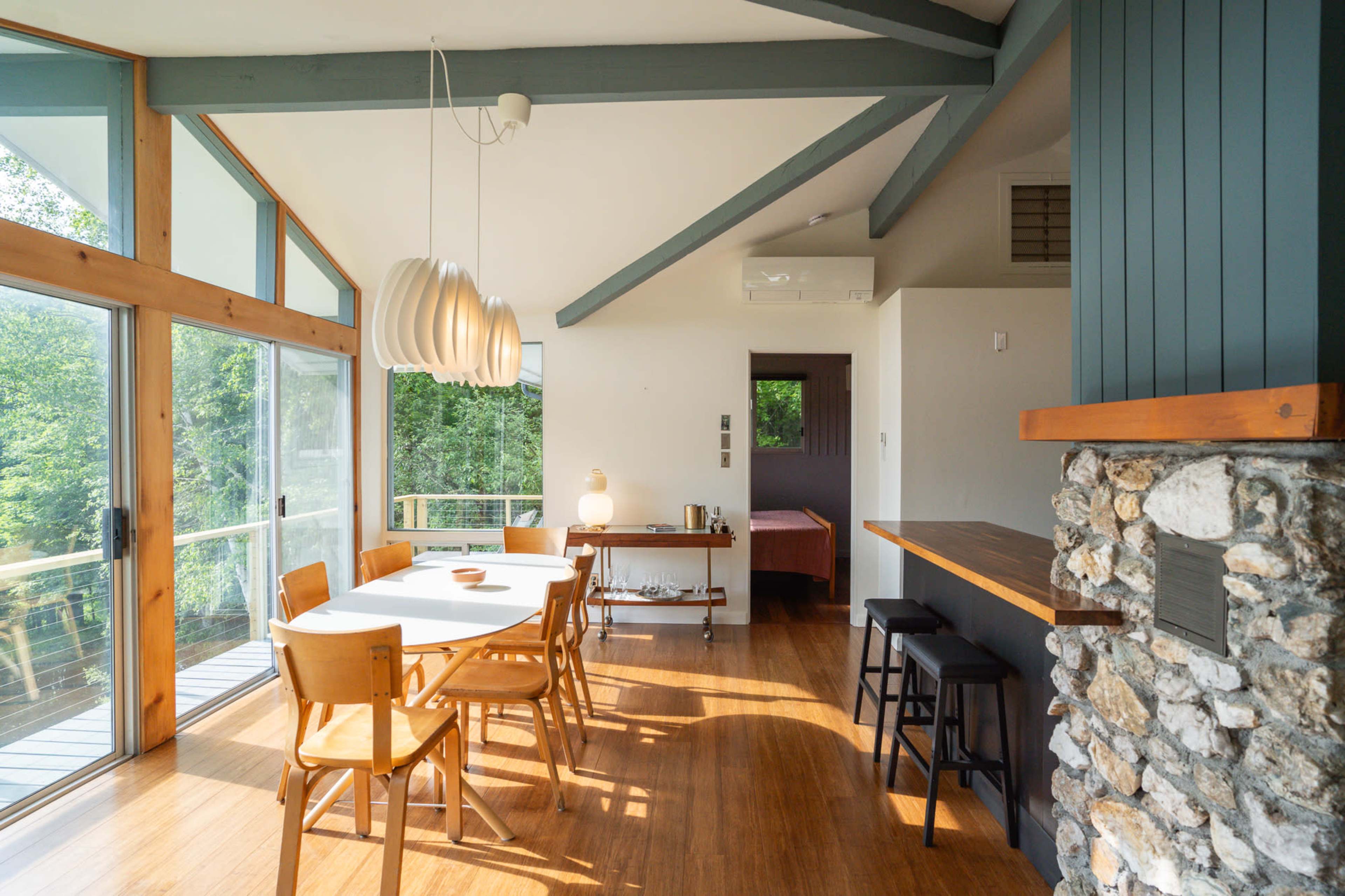 A dining area features a long table surrounded by wooden chairs, with a stone accent wall and large windows overlooking greenery.