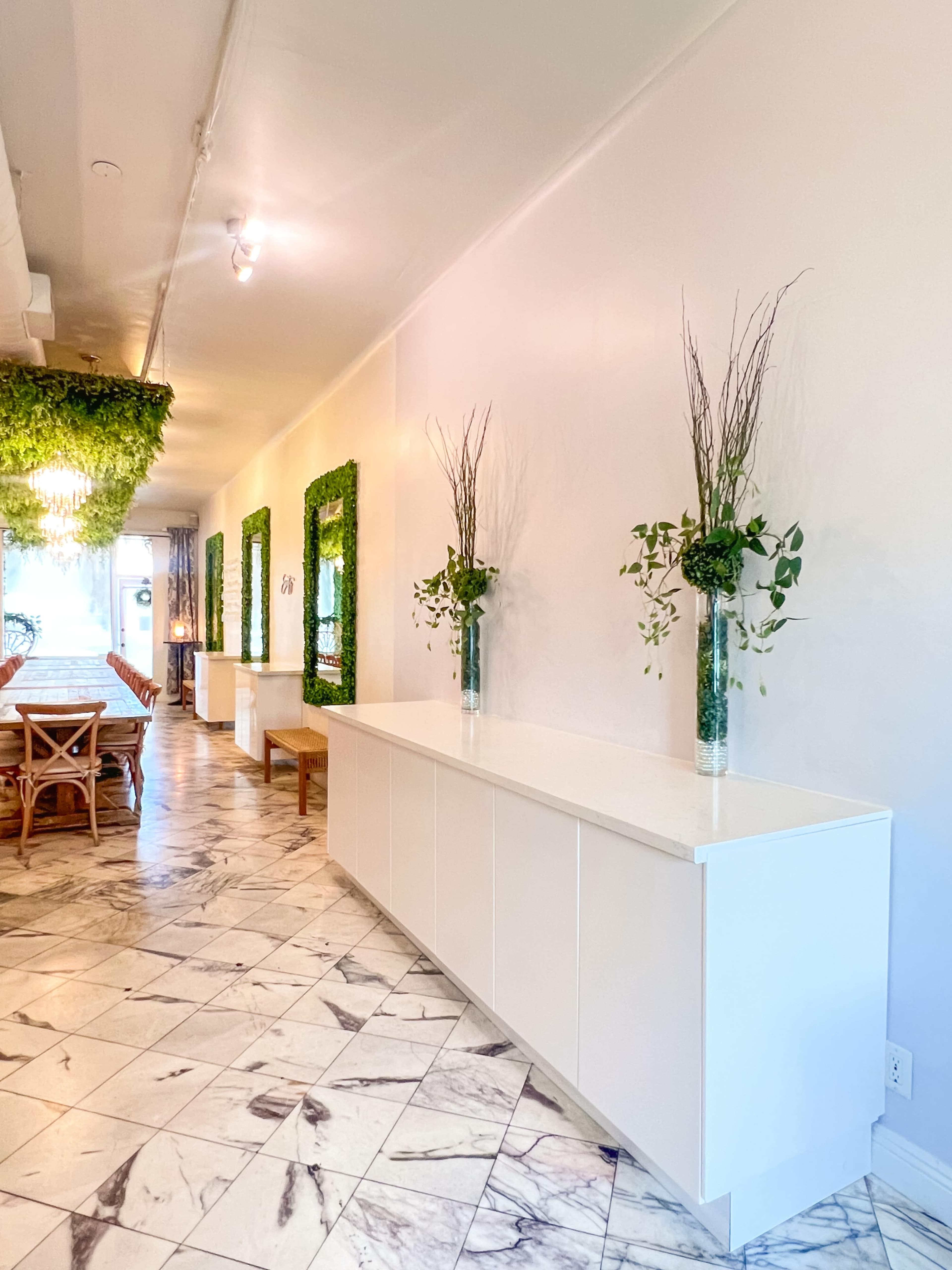 The image shows a hallway with white walls, a marble floor, and a long white cabinet, accented by vases of greenery and framed with vertical plant installations.