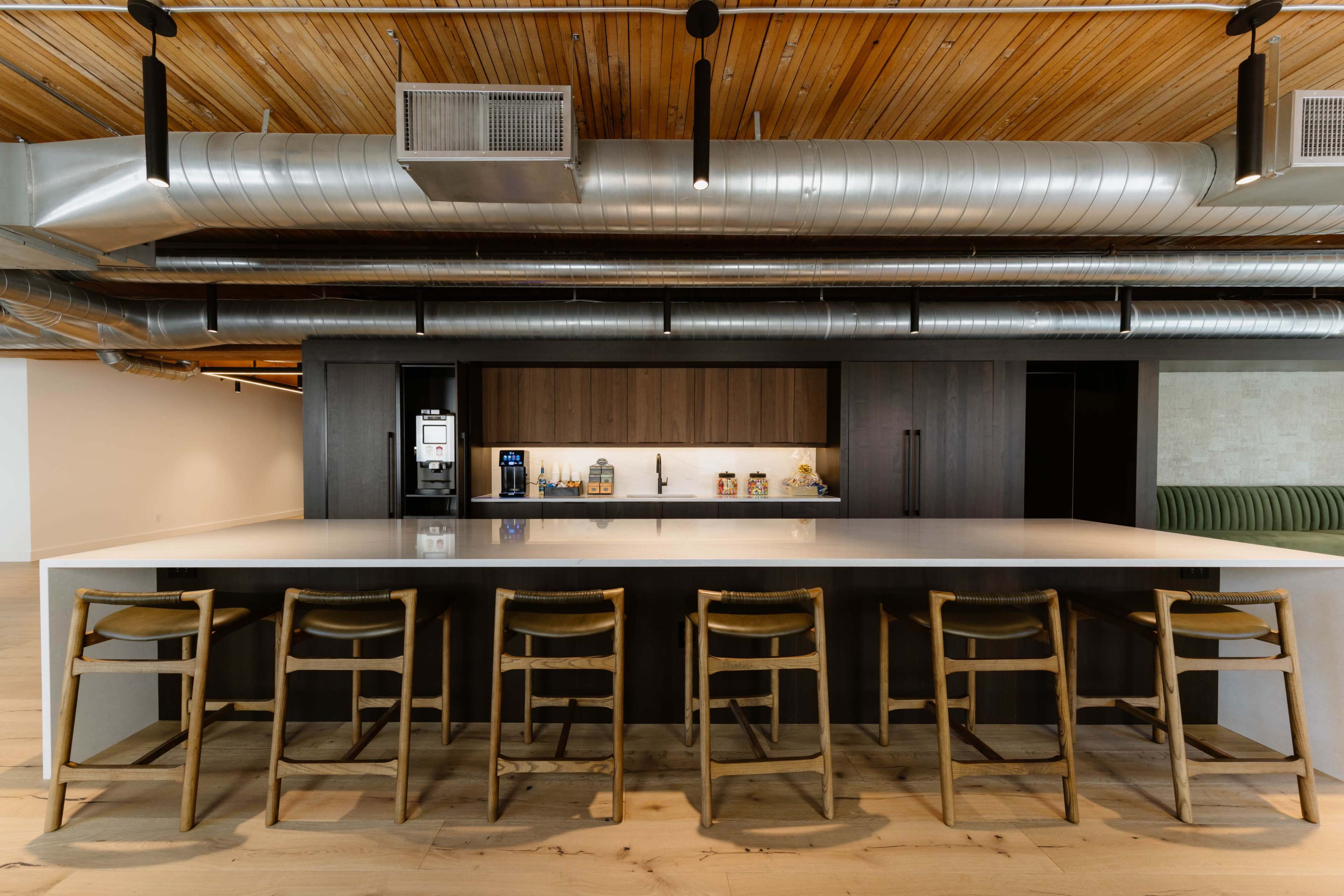 The image shows a modern kitchen with a long, gray countertop and several wooden bar stools, set under a ceiling featuring exposed metal ductwork.