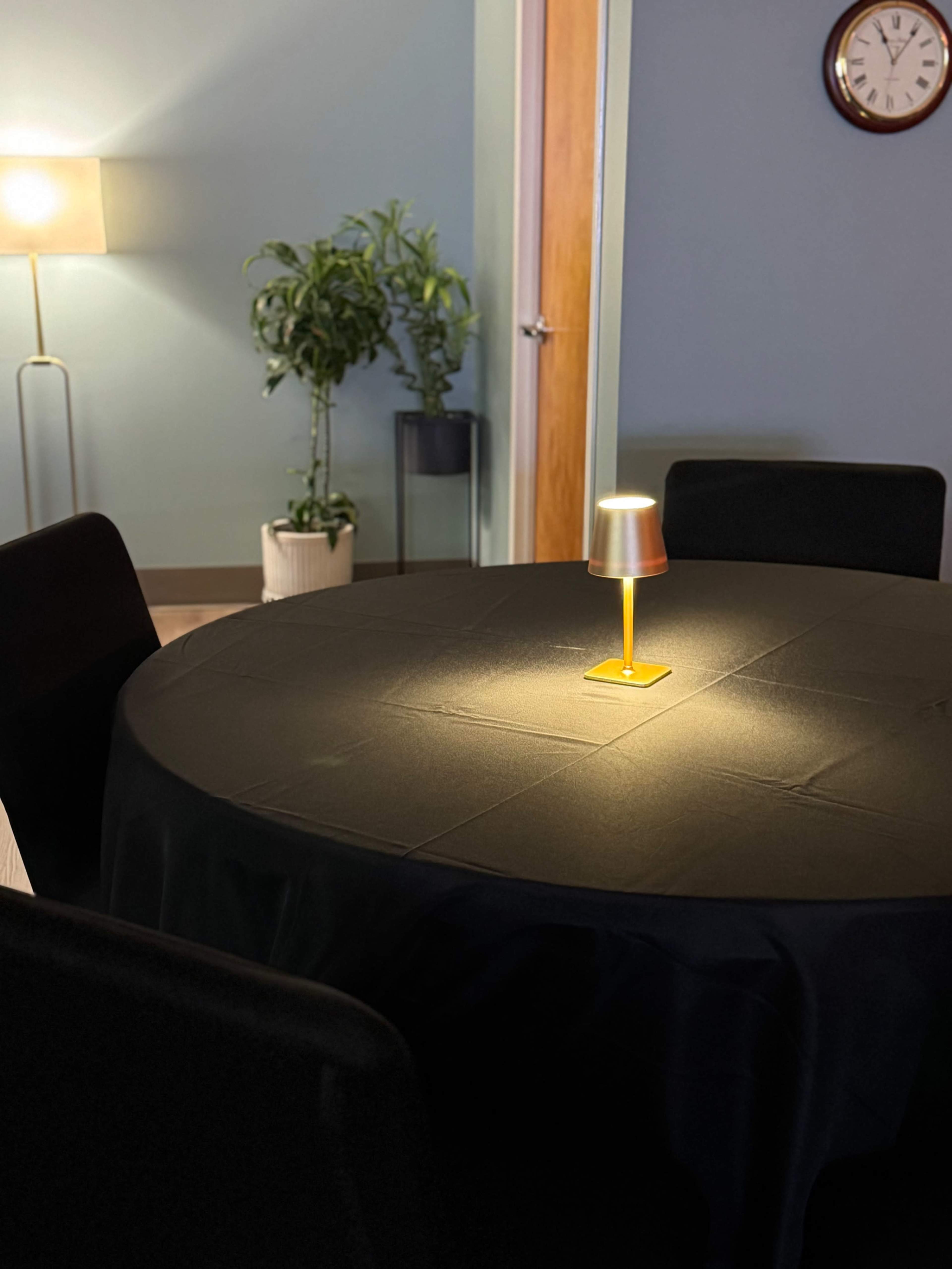 A round table with a black tablecloth is illuminated by a lamp in the center, surrounded by four chairs in a softly lit room featuring a plant and a wall clock.