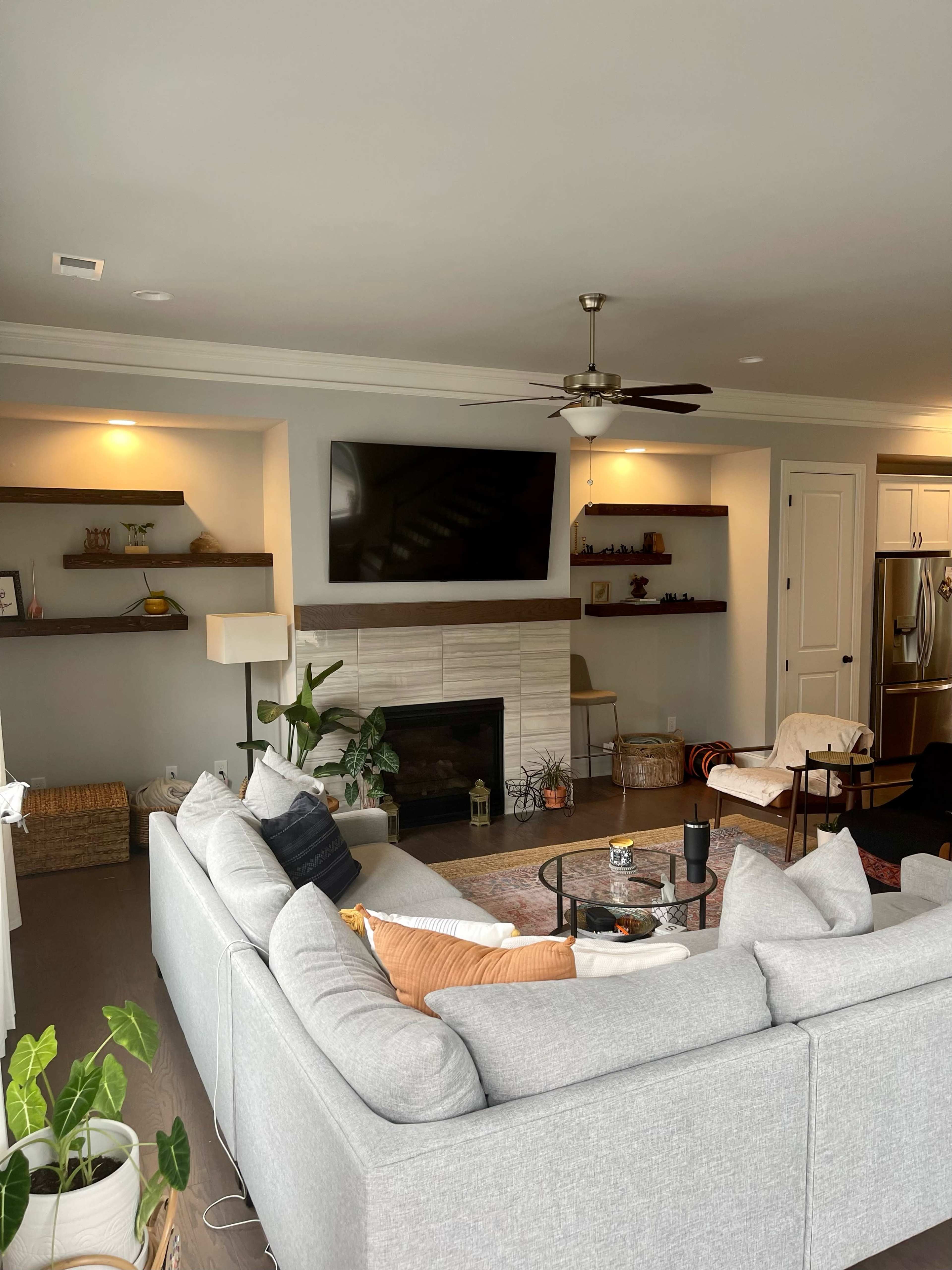 A modern living room with a gray sectional sofa, a fireplace with a TV mounted above it, and floating shelves on either side, featuring decorative items and plants.