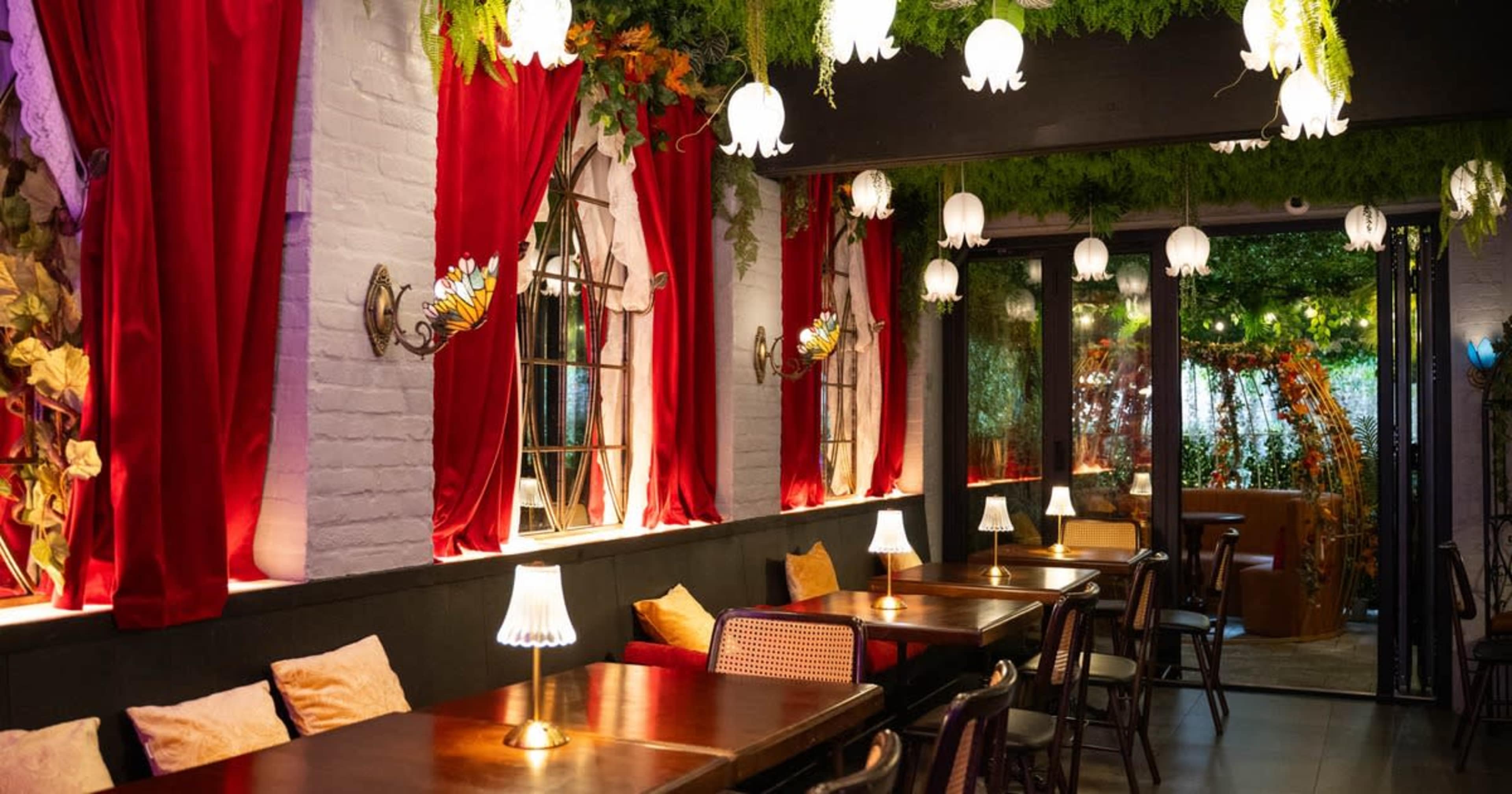 The image shows a restaurant interior with wooden tables, soft lighting from decorative lamps, and walls adorned with greenery and red curtains.