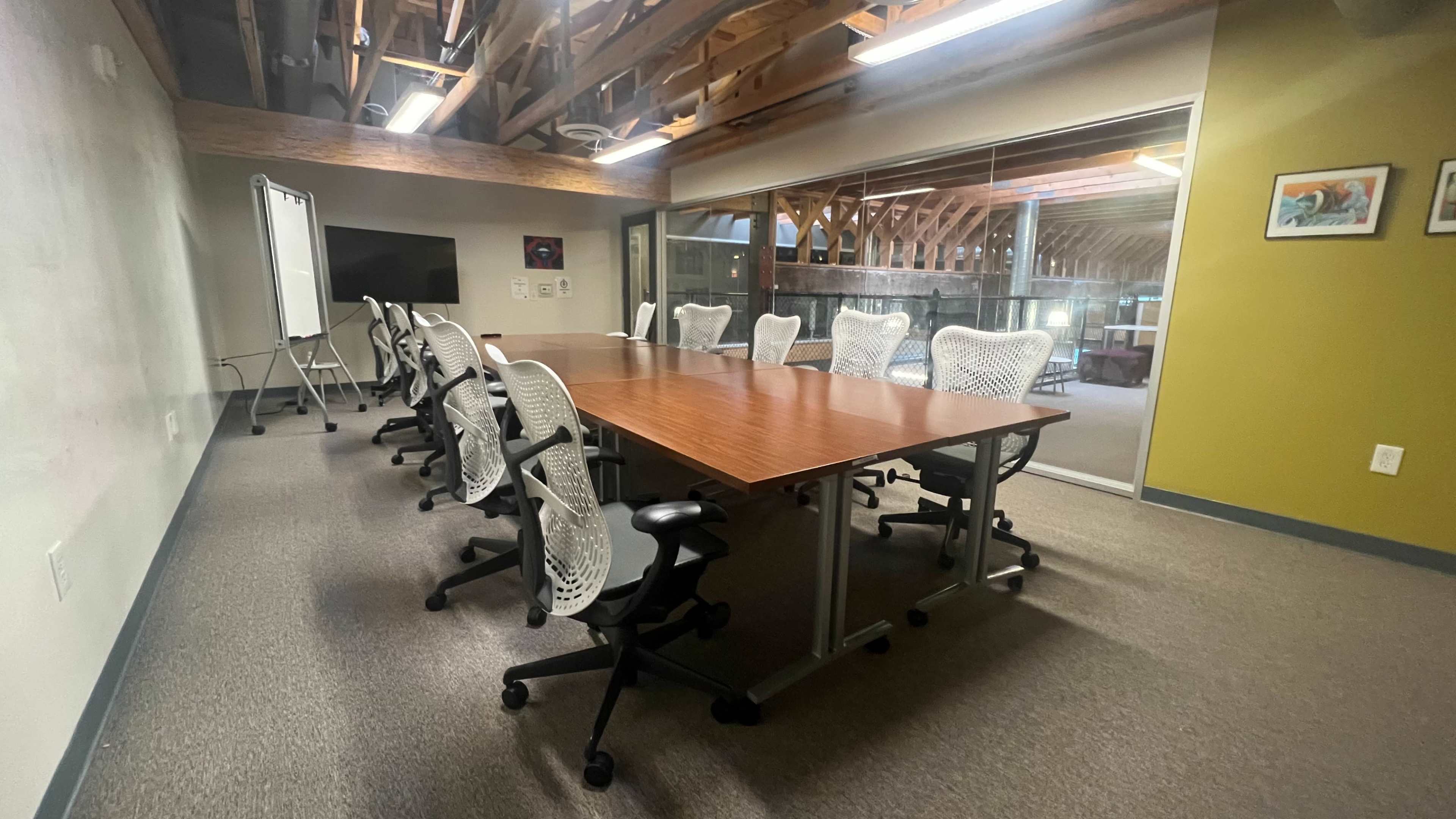 A spacious conference room features a large wooden table surrounded by ergonomic chairs, with large windows revealing a view of a wooden-beamed structure.