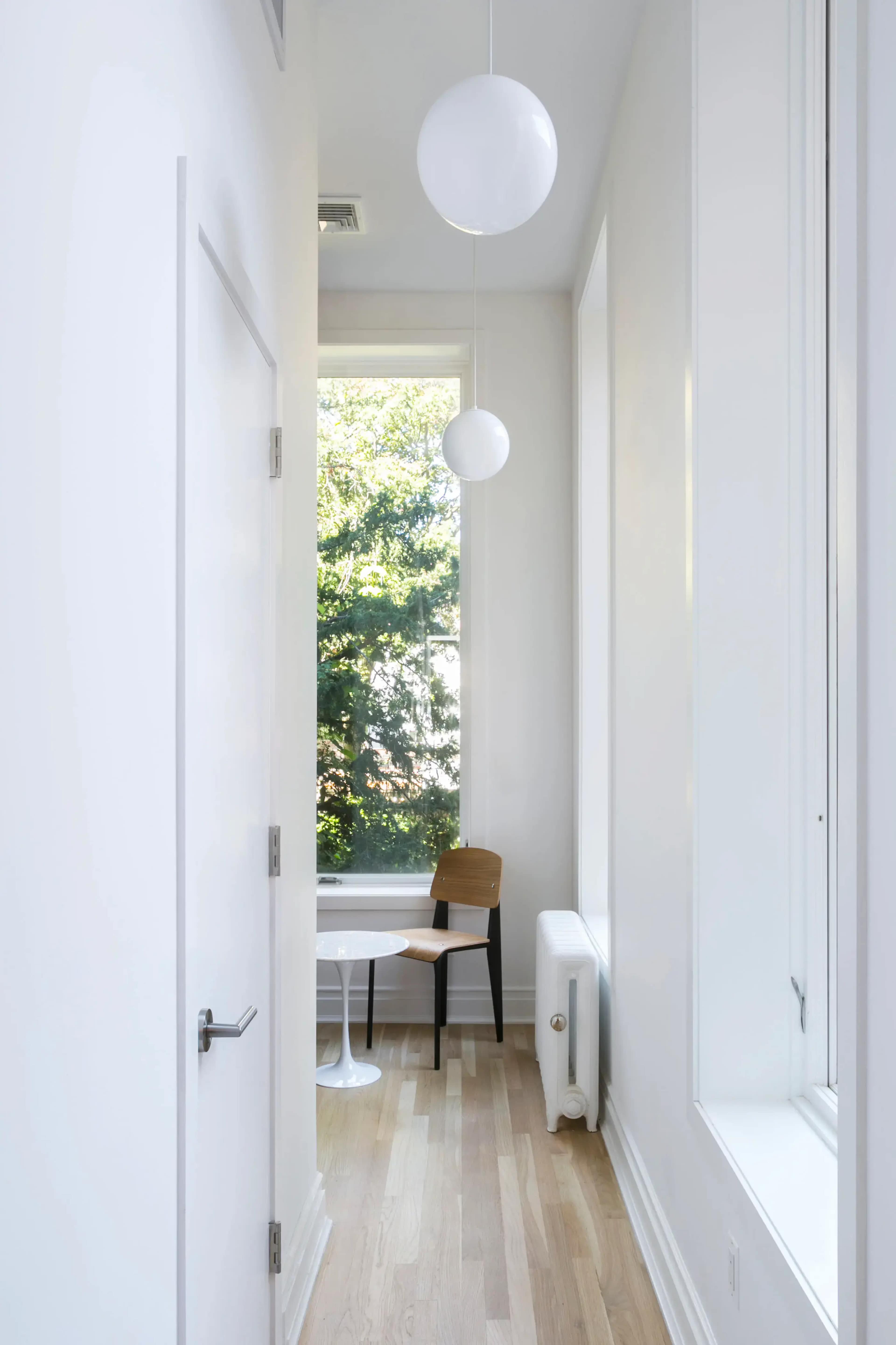 A narrow hallway features a white door, a window with a view of greenery outside, a round table, a chair, and two pendant lights overhead.