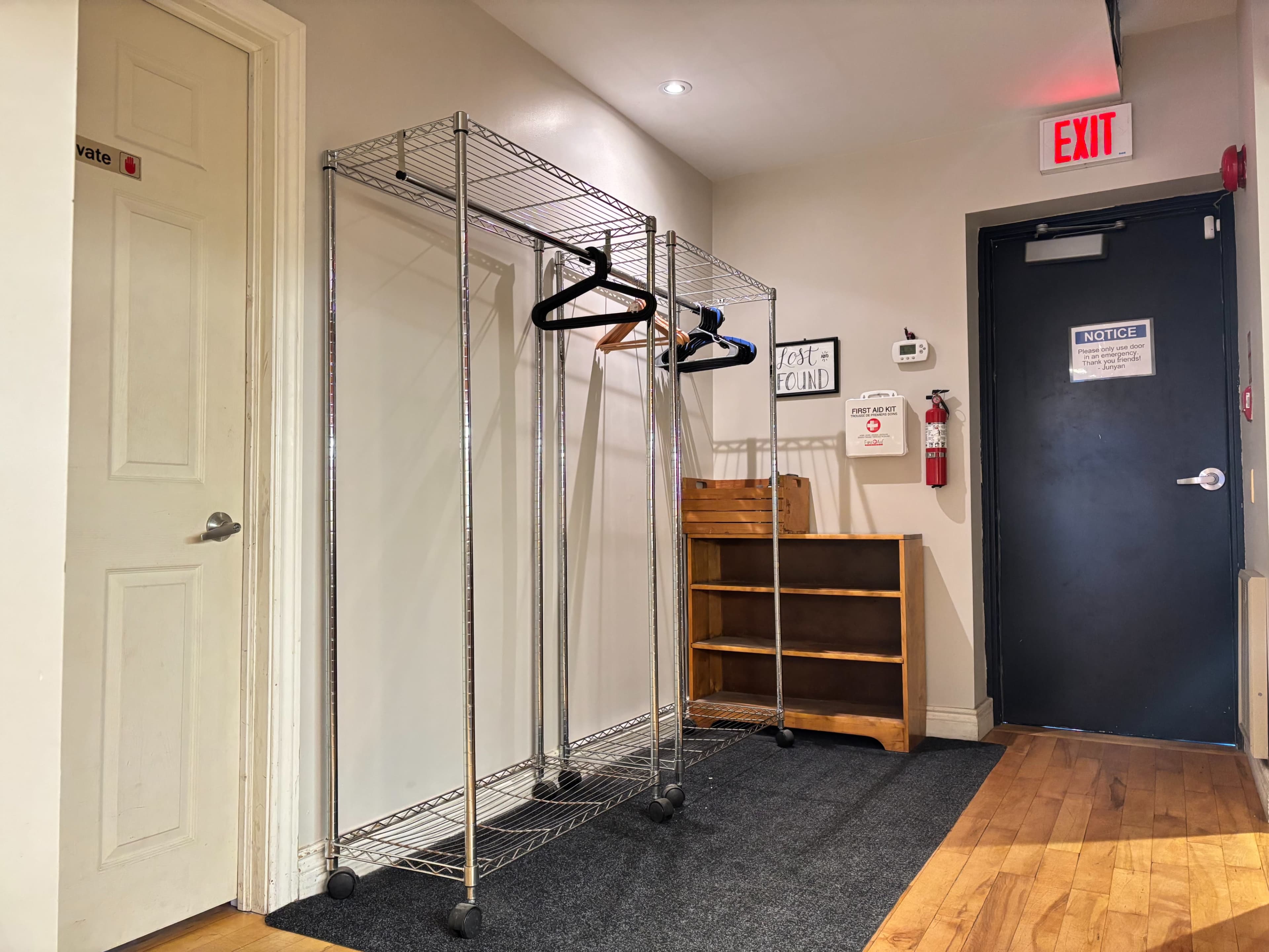 The image shows a small hallway area with two clothing racks on one side and a wooden shelf on the opposite side, next to a door marked with an exit sign.
