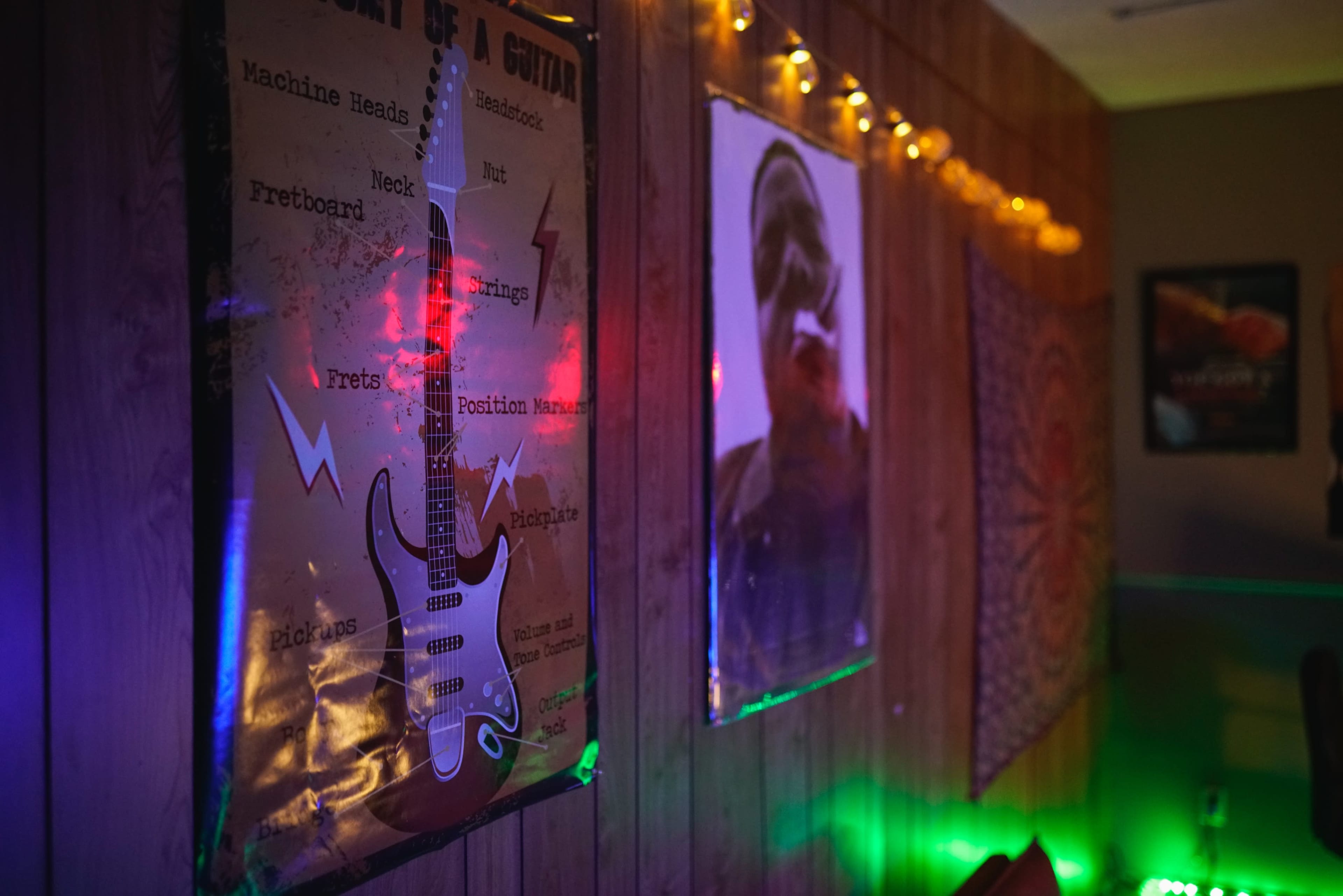 The image shows a room with wooden walls featuring posters of a guitar anatomy diagram and a person, illuminated by soft colored lights and strings of fairy lights.