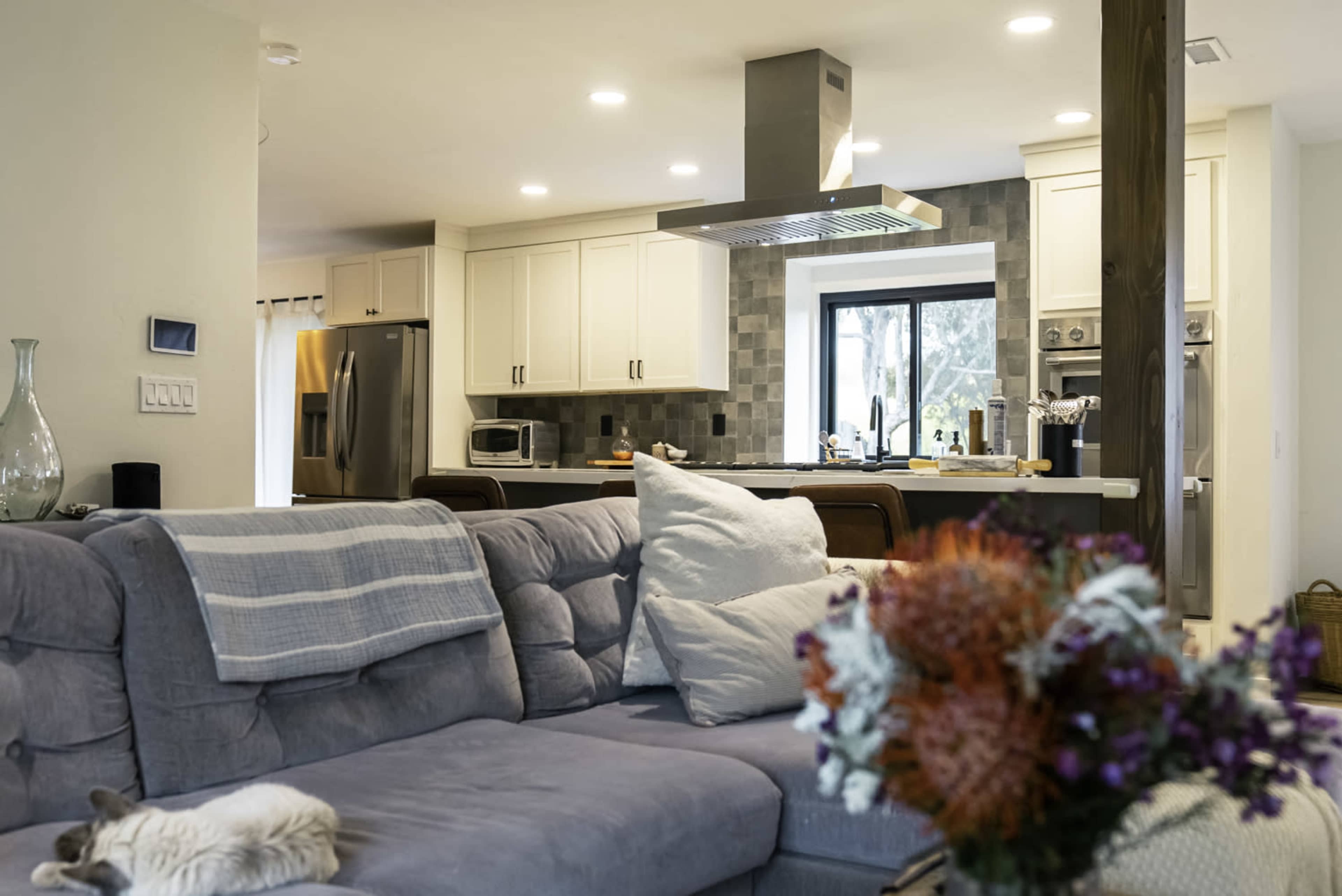 A modern kitchen is visible in the background, featuring stainless steel appliances and white cabinetry, with a cozy living area in the foreground that includes a gray couch and decorative flowers.