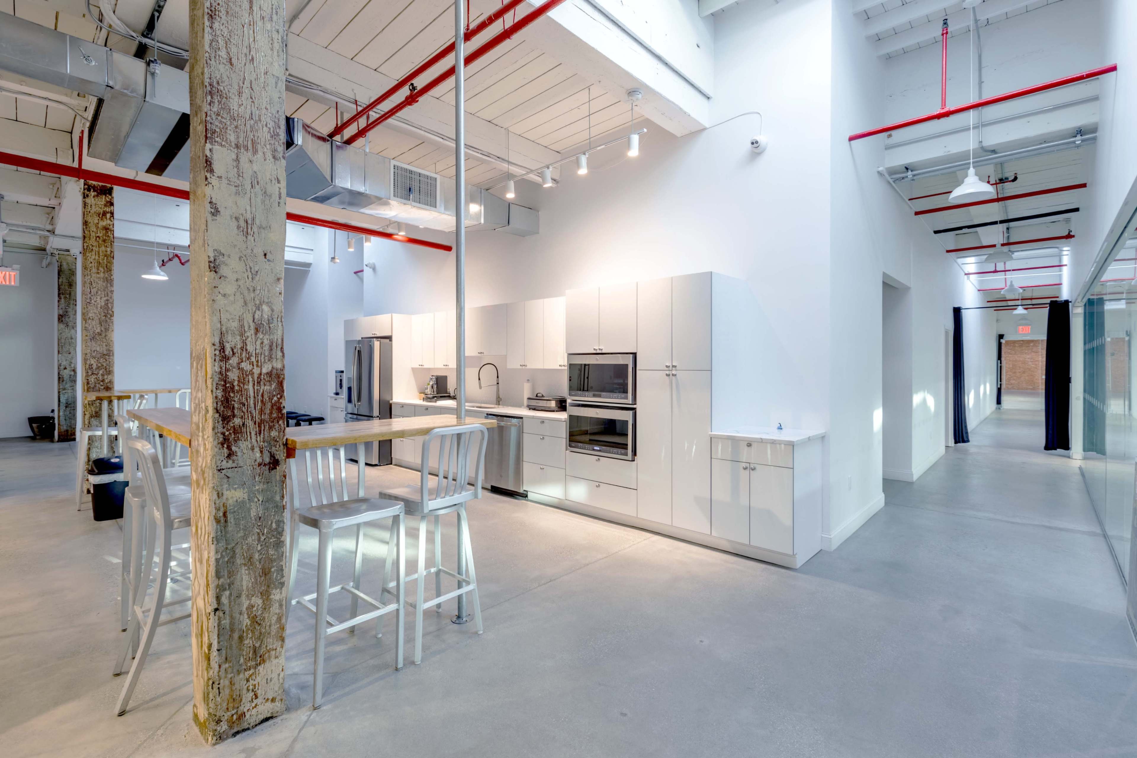 The image depicts a modern, minimalist kitchen area with white cabinets and appliances, adjacent to a spacious corridor in a well-lit industrial-style interior.