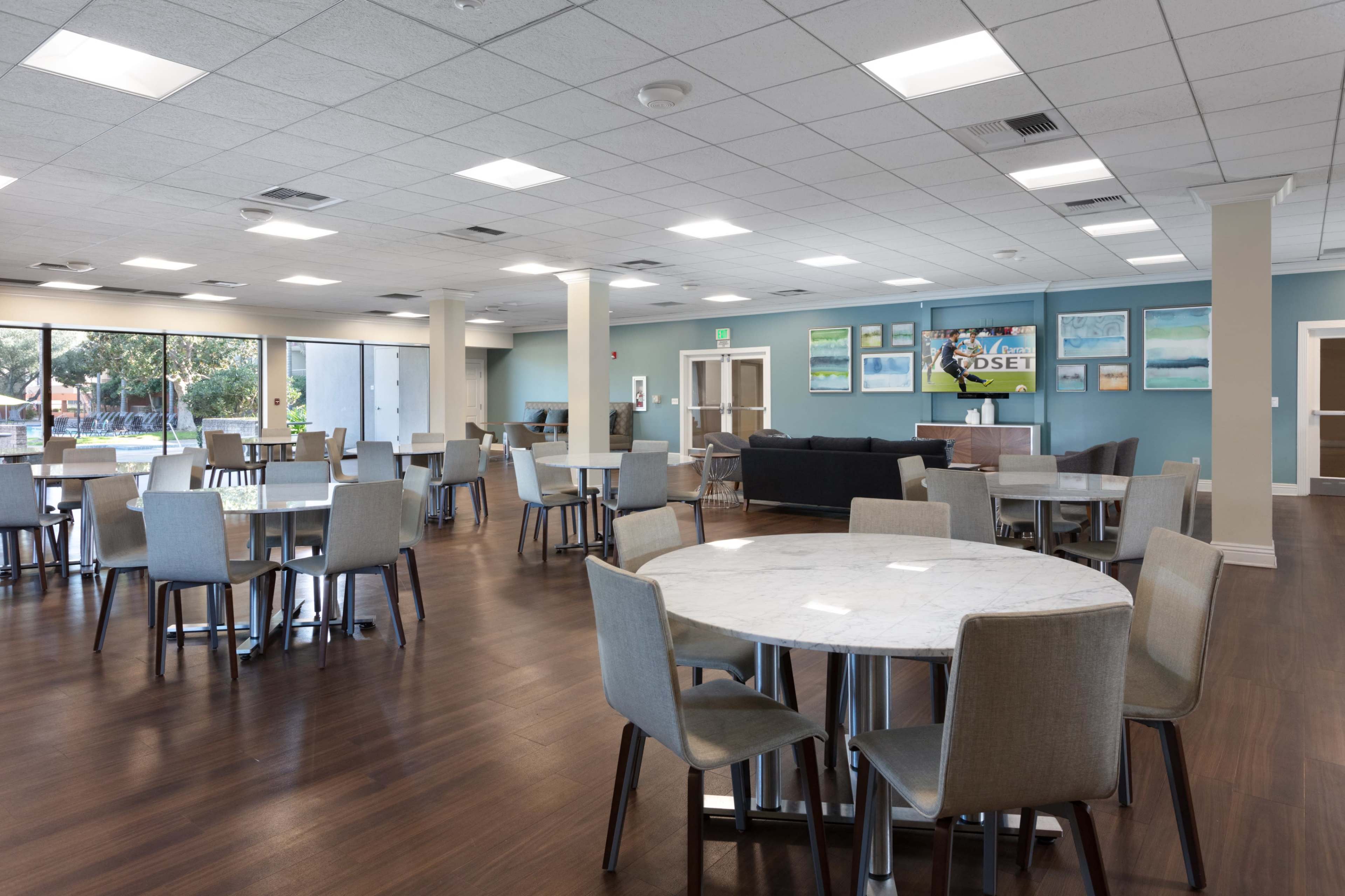 The image shows a spacious and modern communal dining area with multiple tables and chairs arranged for seating, alongside large windows that allow natural light to fill the room.