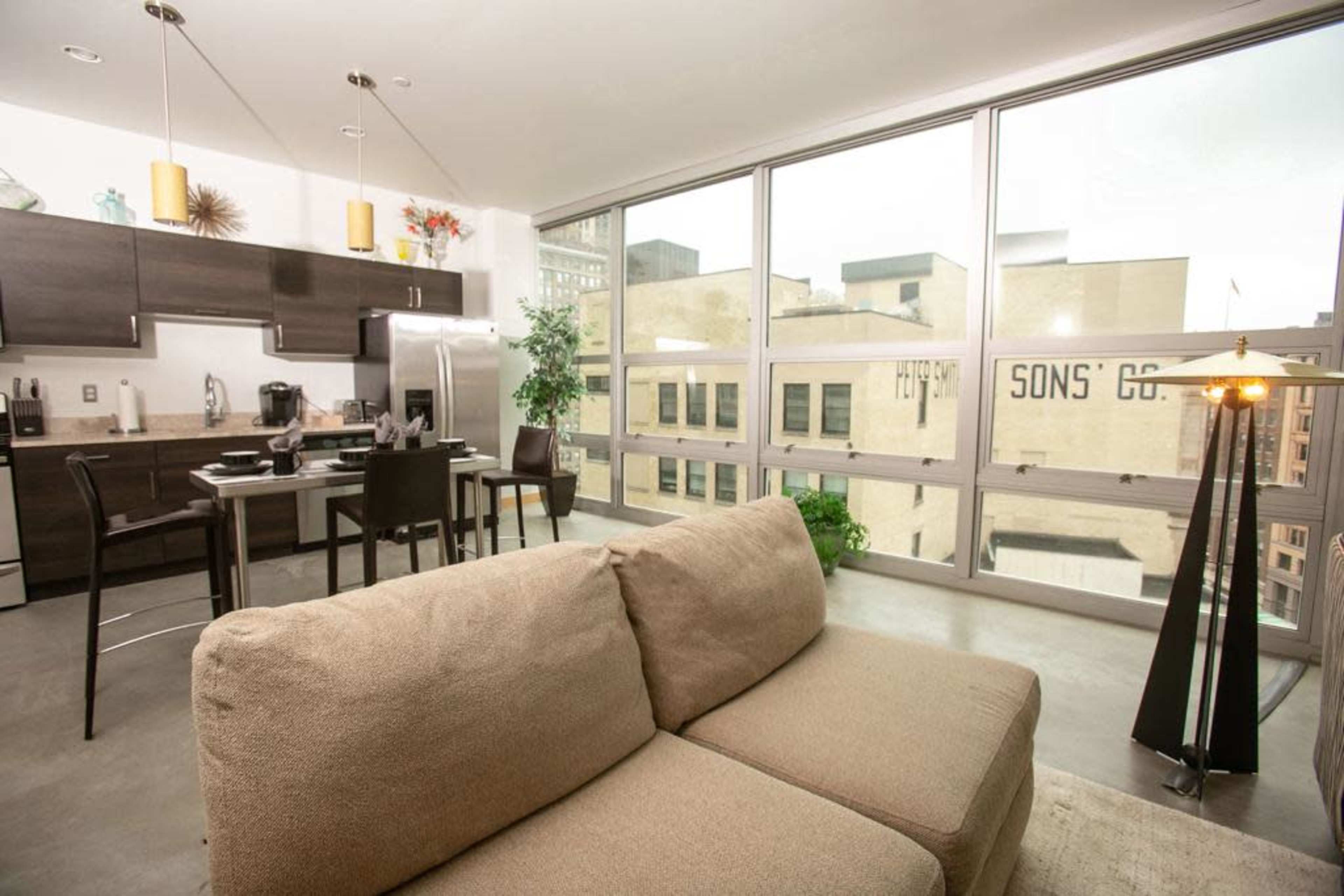 A modern kitchen and dining area are visible, featuring a large window overlooking a cityscape and a comfortable sofa in the foreground.