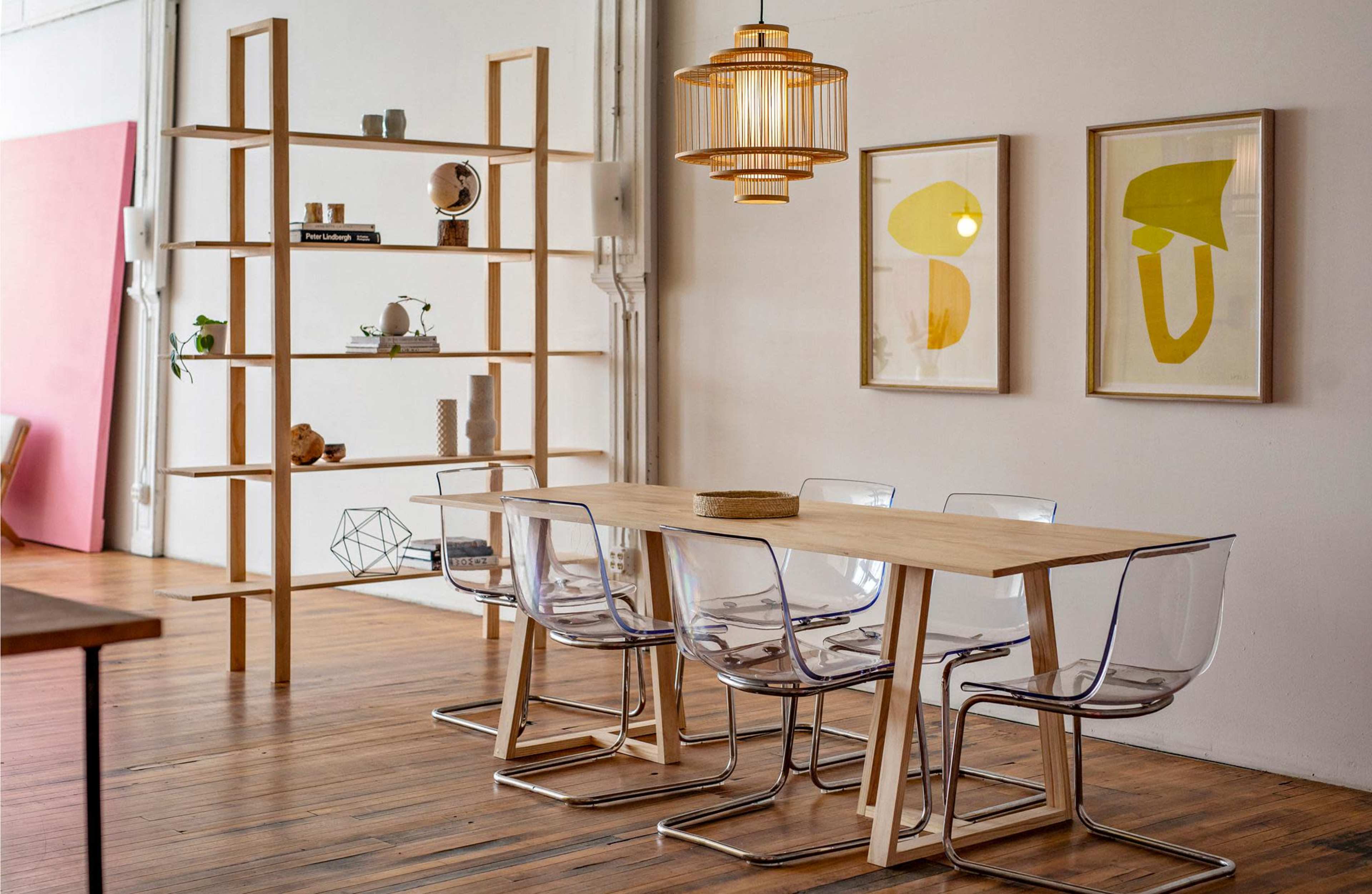 A modern dining area features a wooden table, clear acrylic chairs, a tall wooden shelf, and framed artworks on the wall.