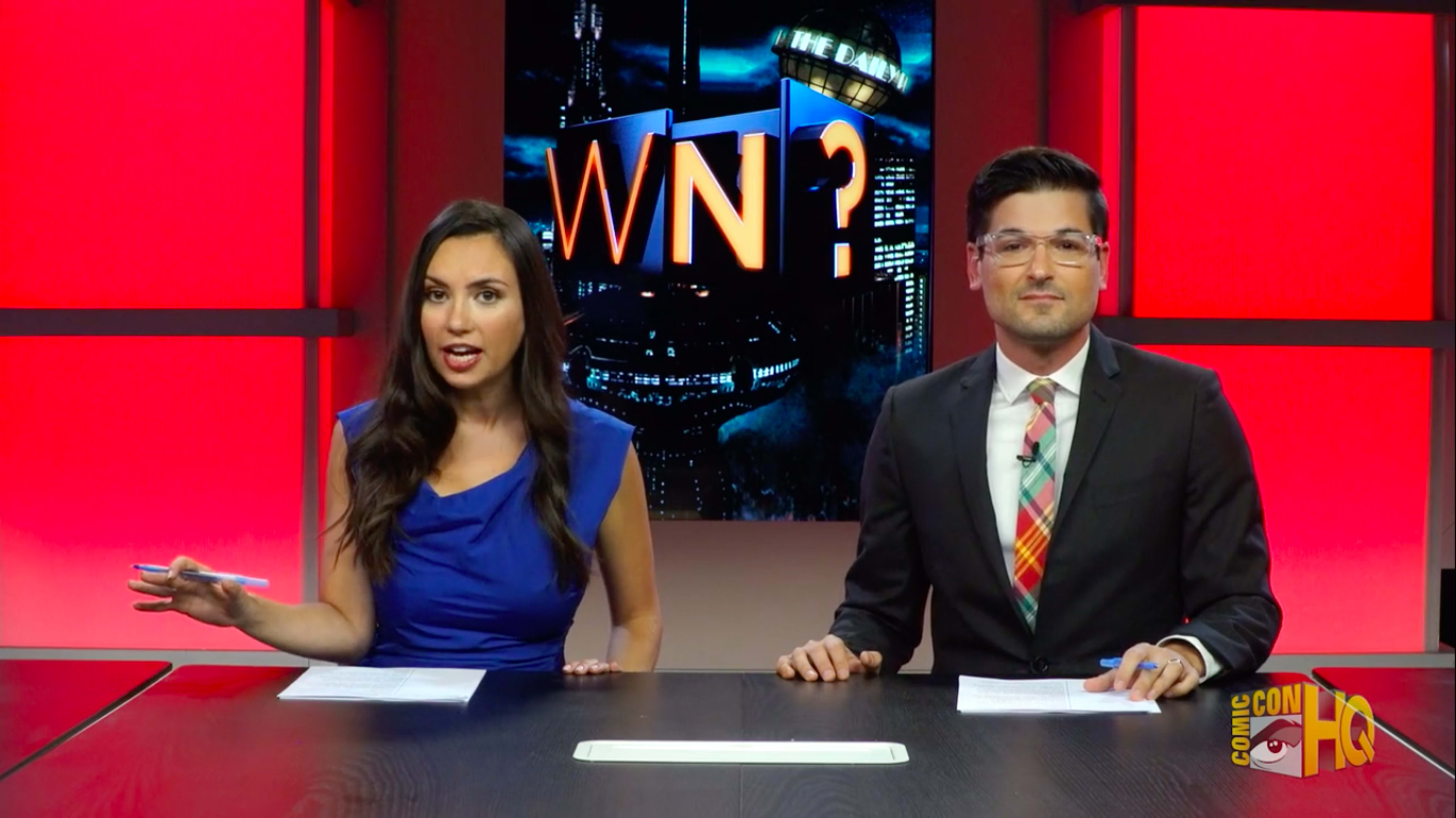 Two hosts are seated at a table in a studio with a brightly lit backdrop displaying "WN?" and a city skyline.