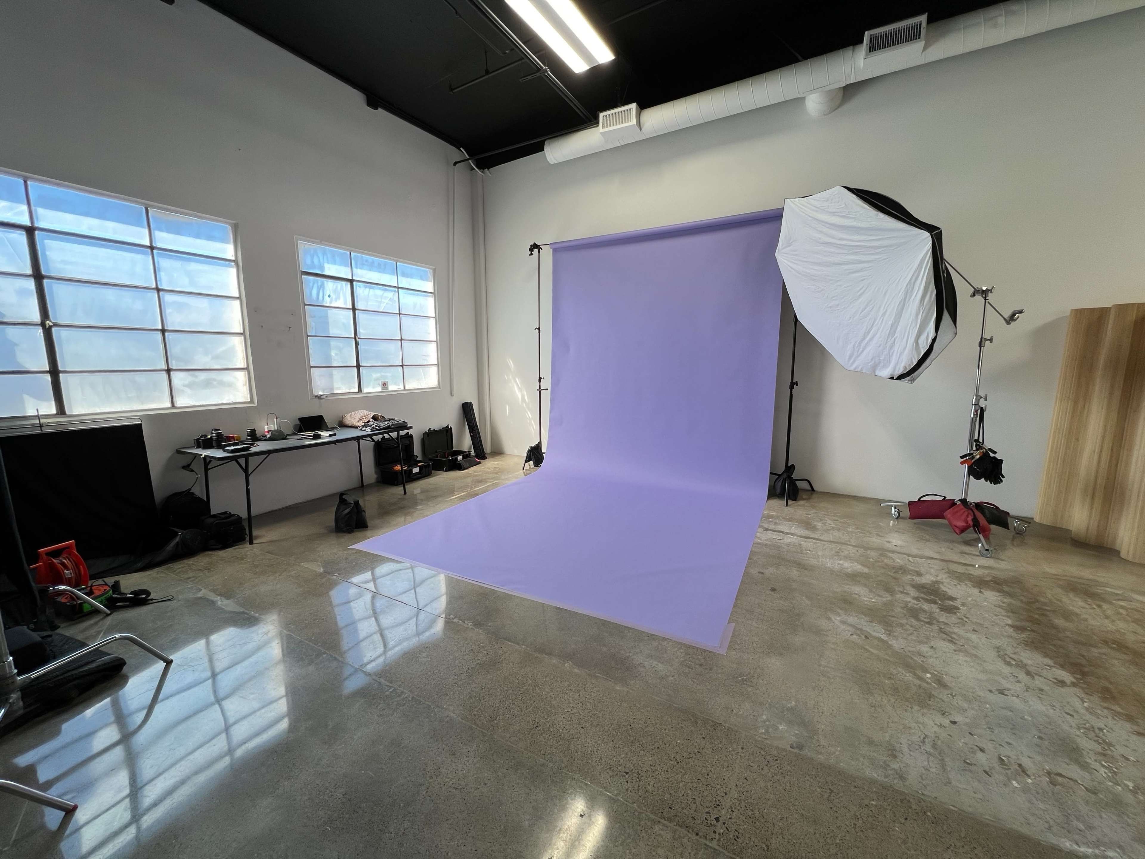 A photography studio features a lavender backdrop on a polished concrete floor, with large windows illuminating the space and various equipment visible in the background.