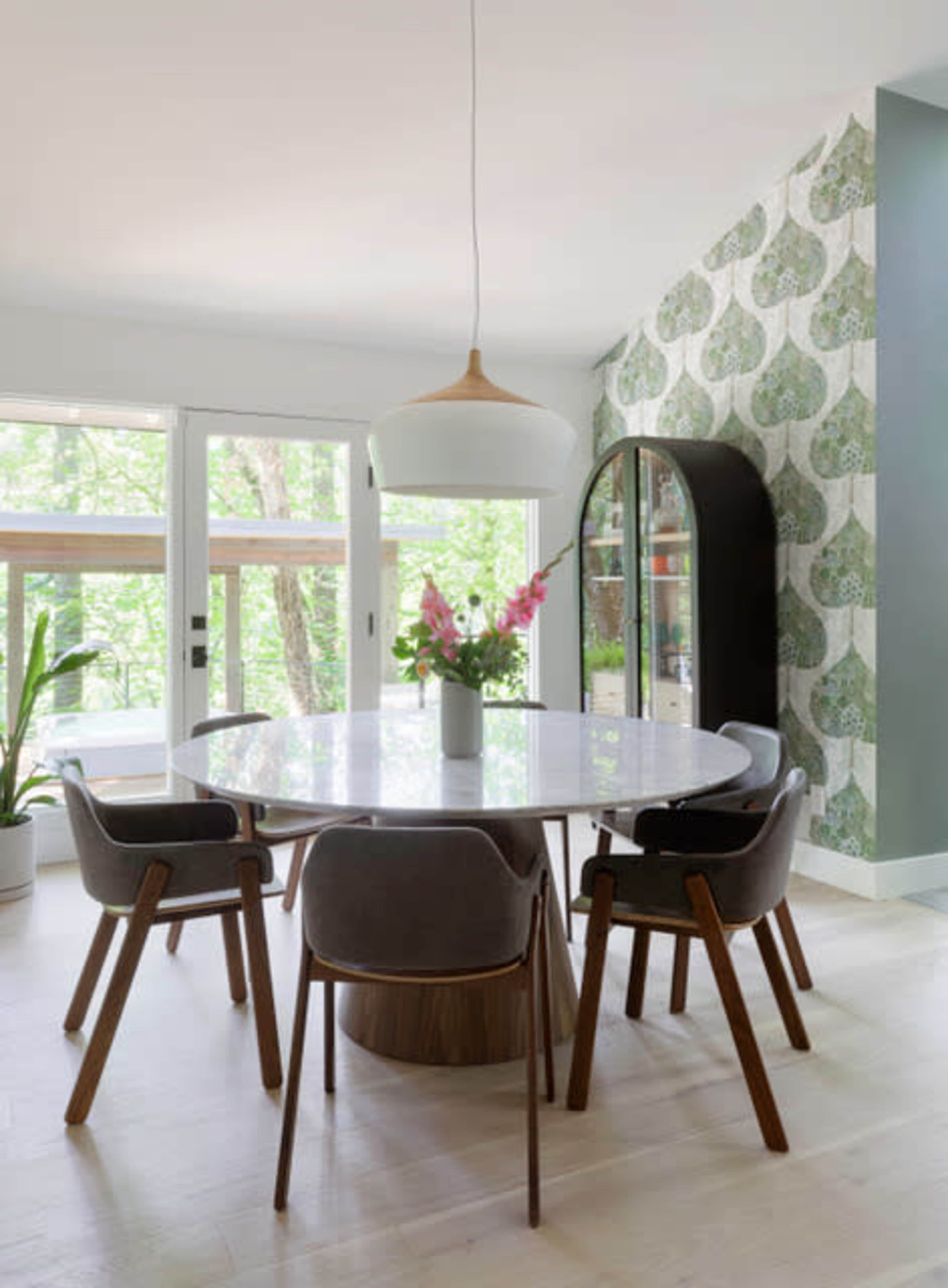 A round dining table with four chairs is set in a light-filled room featuring a window view of greenery and a patterned wallpaper.