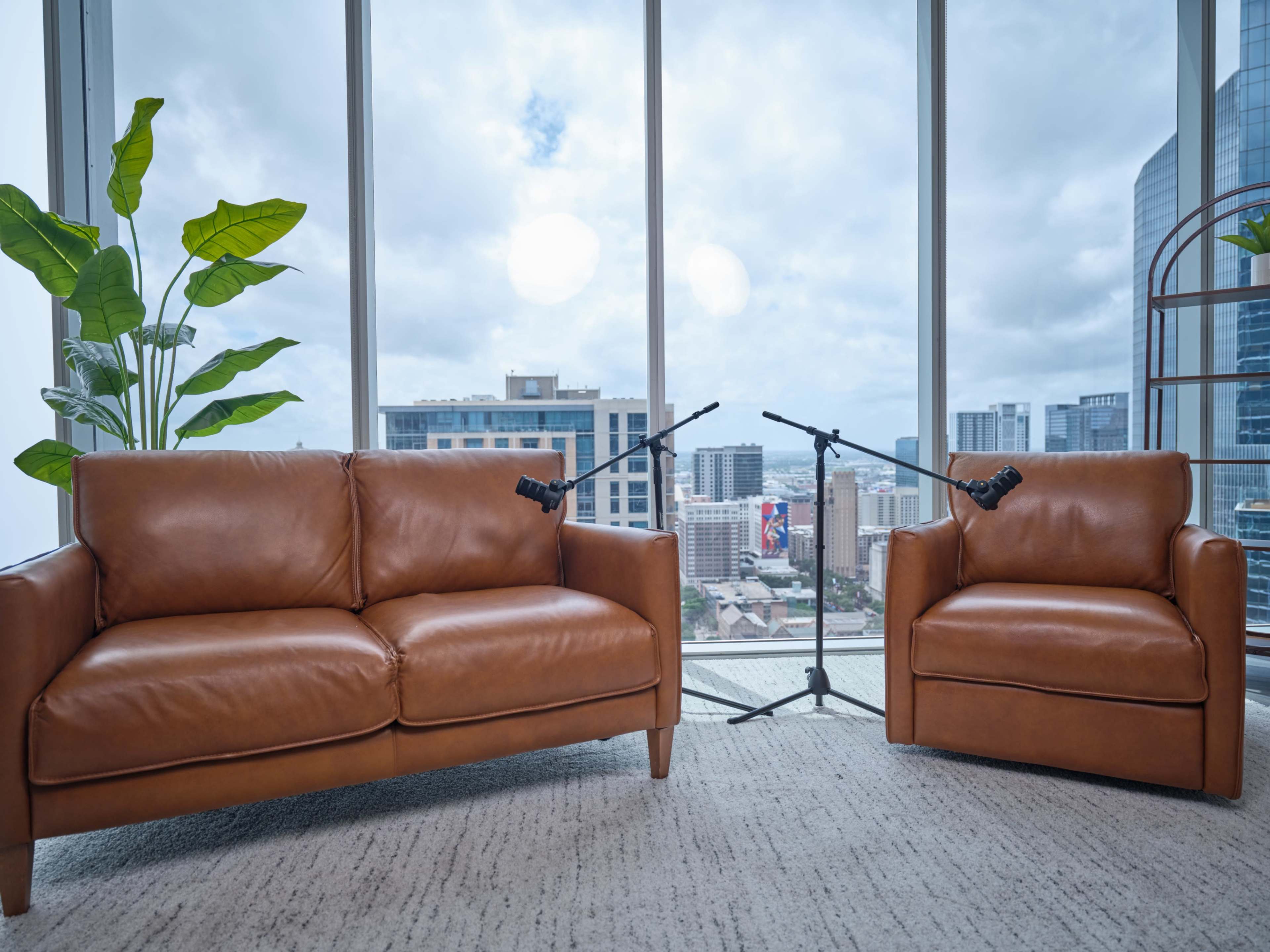 The image shows two brown leather sofas positioned in front of large windows with a city view, accompanied by two microphones on stands.