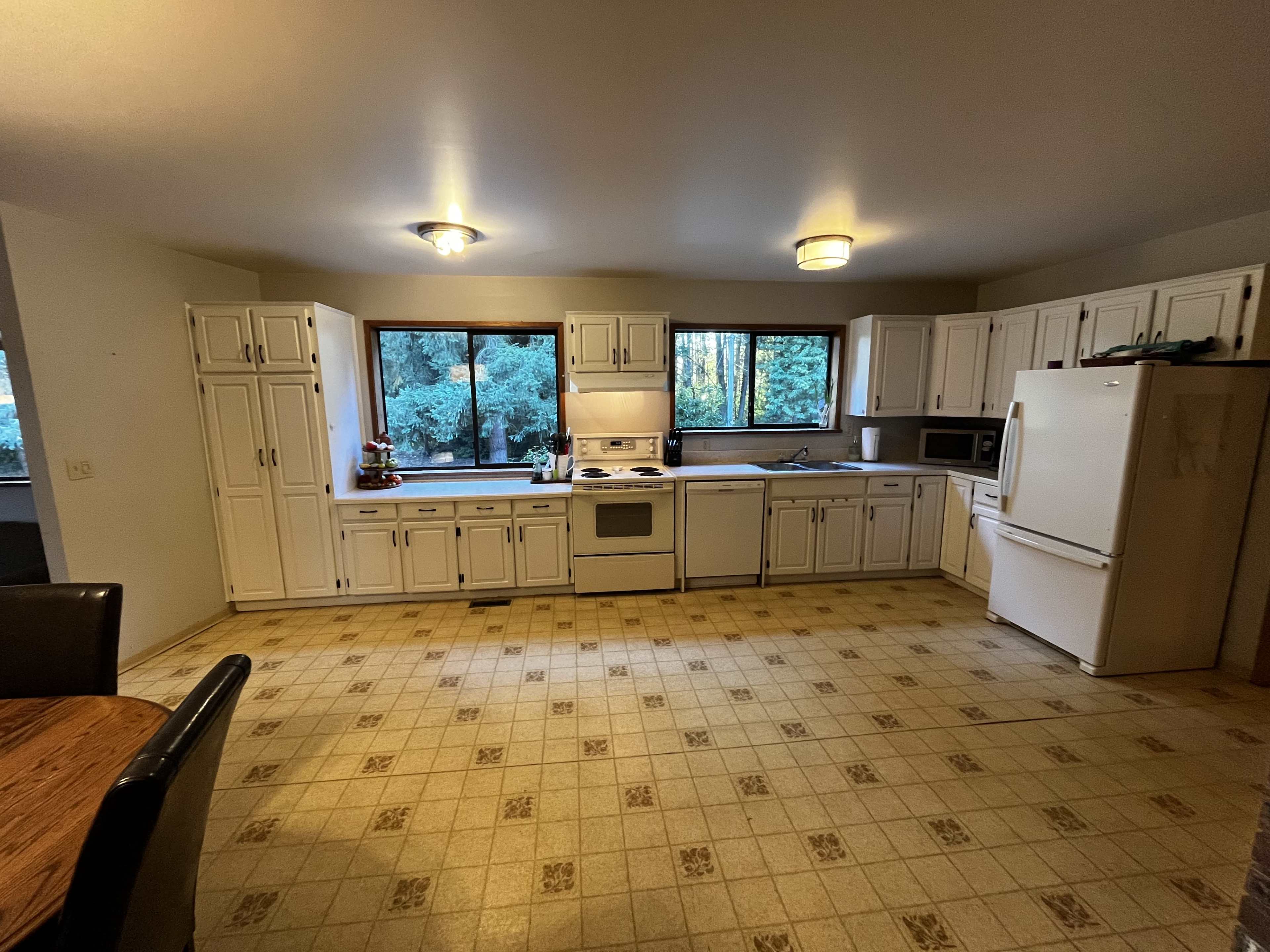 A spacious kitchen features white cabinetry, a stove, an oven, a microwave, and large windows overlooking a wooded area.