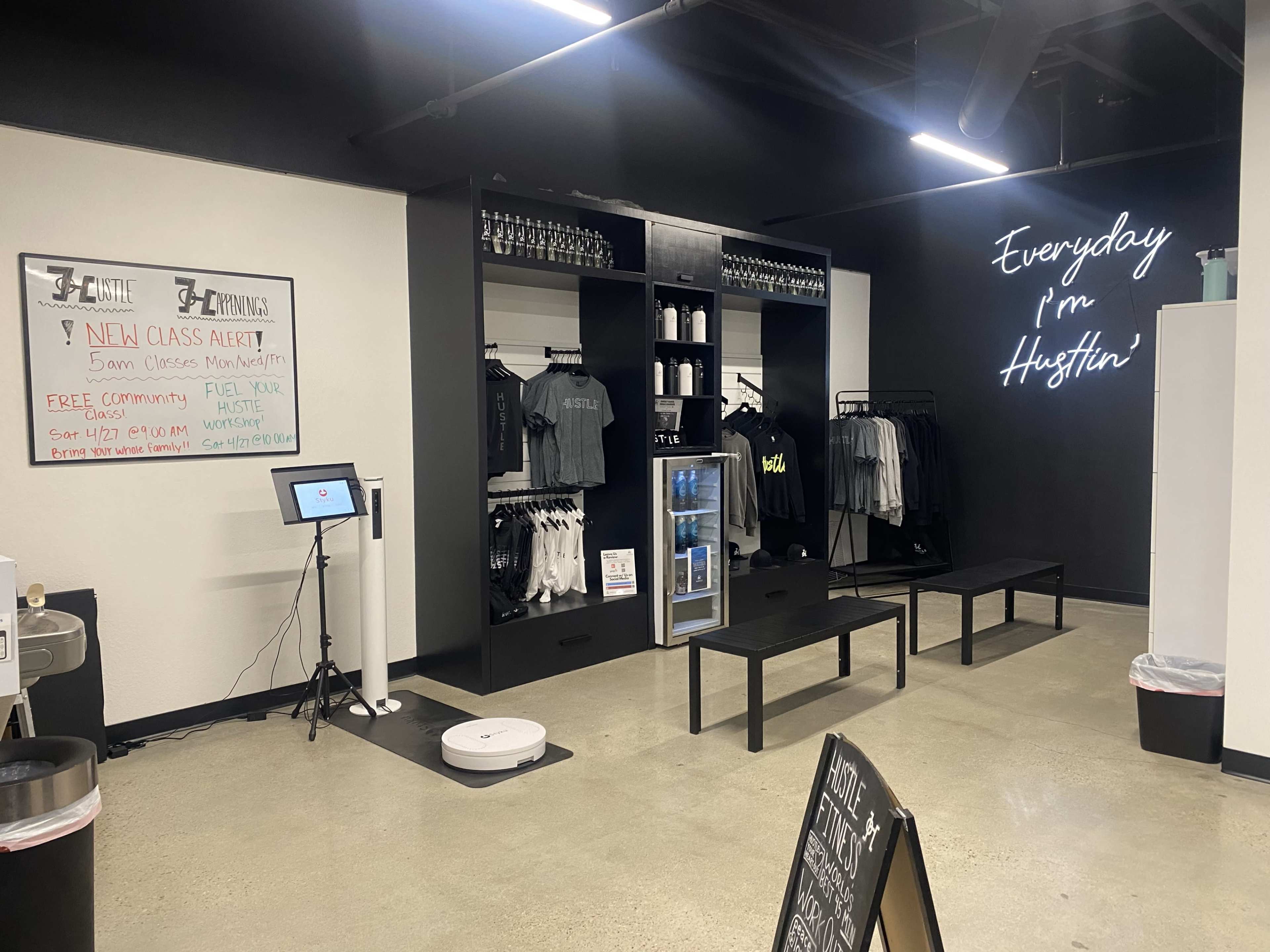 The interior of a fitness studio features black walls, shelves with apparel, a sign displaying "Everyday I'm Hustlin'," and a whiteboard announcing new classes.