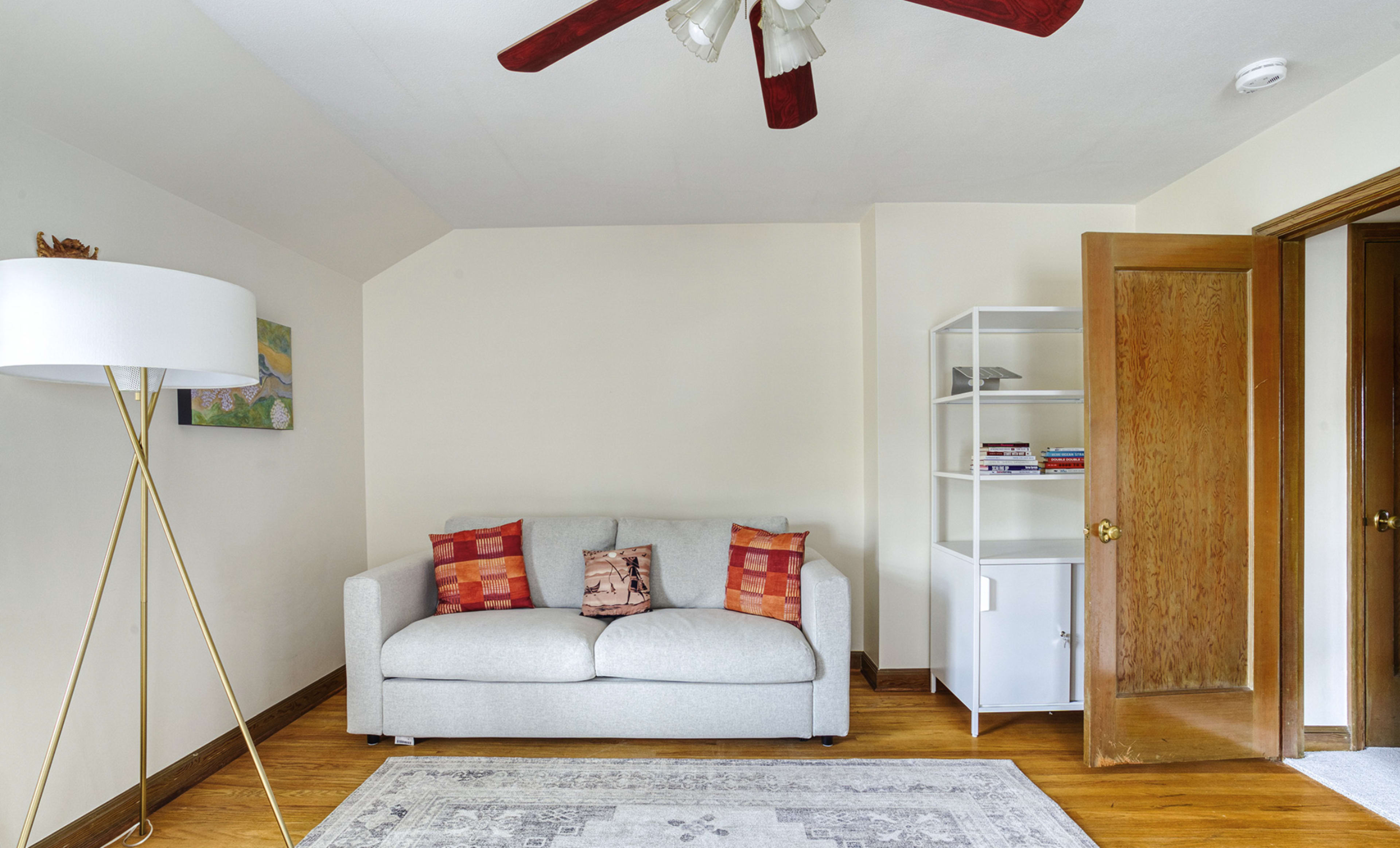 The image depicts a small, well-lit living room featuring a light gray sofa with decorative pillows, a floor lamp, a bookshelf, and a closed door leading to another room.
