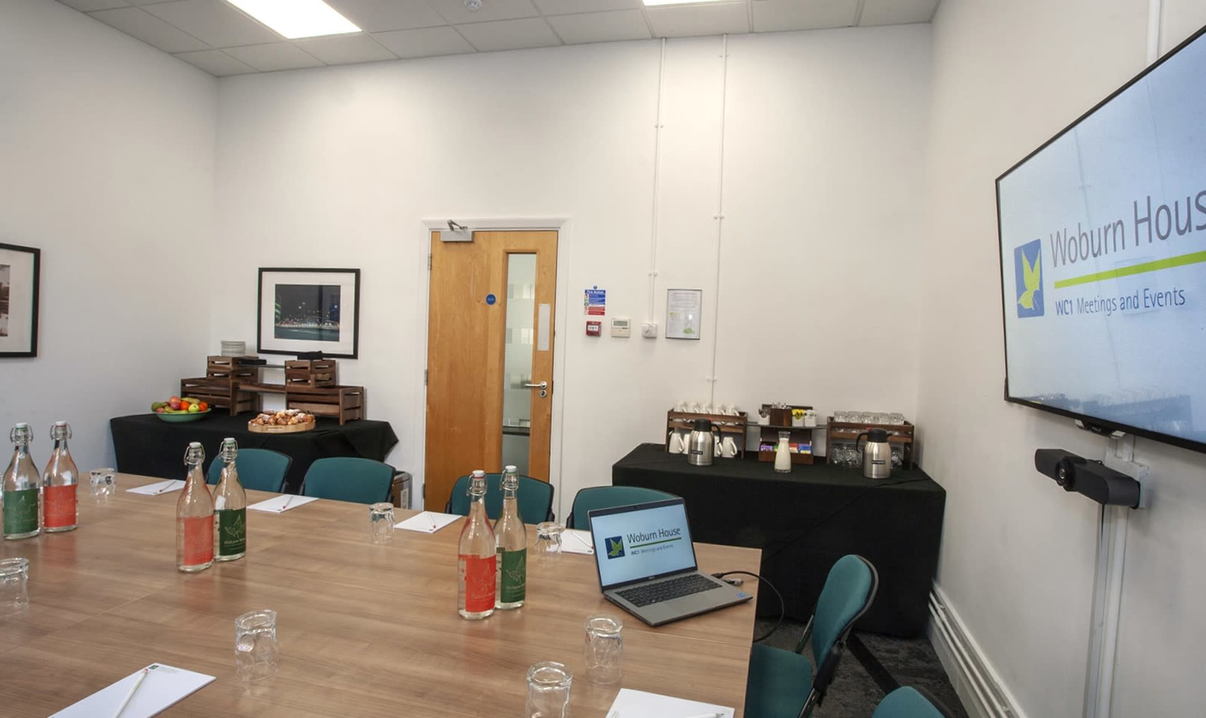 A conference room is set up with a wooden table, chairs, and presentation equipment, featuring a side table with refreshments and an entrance door.
