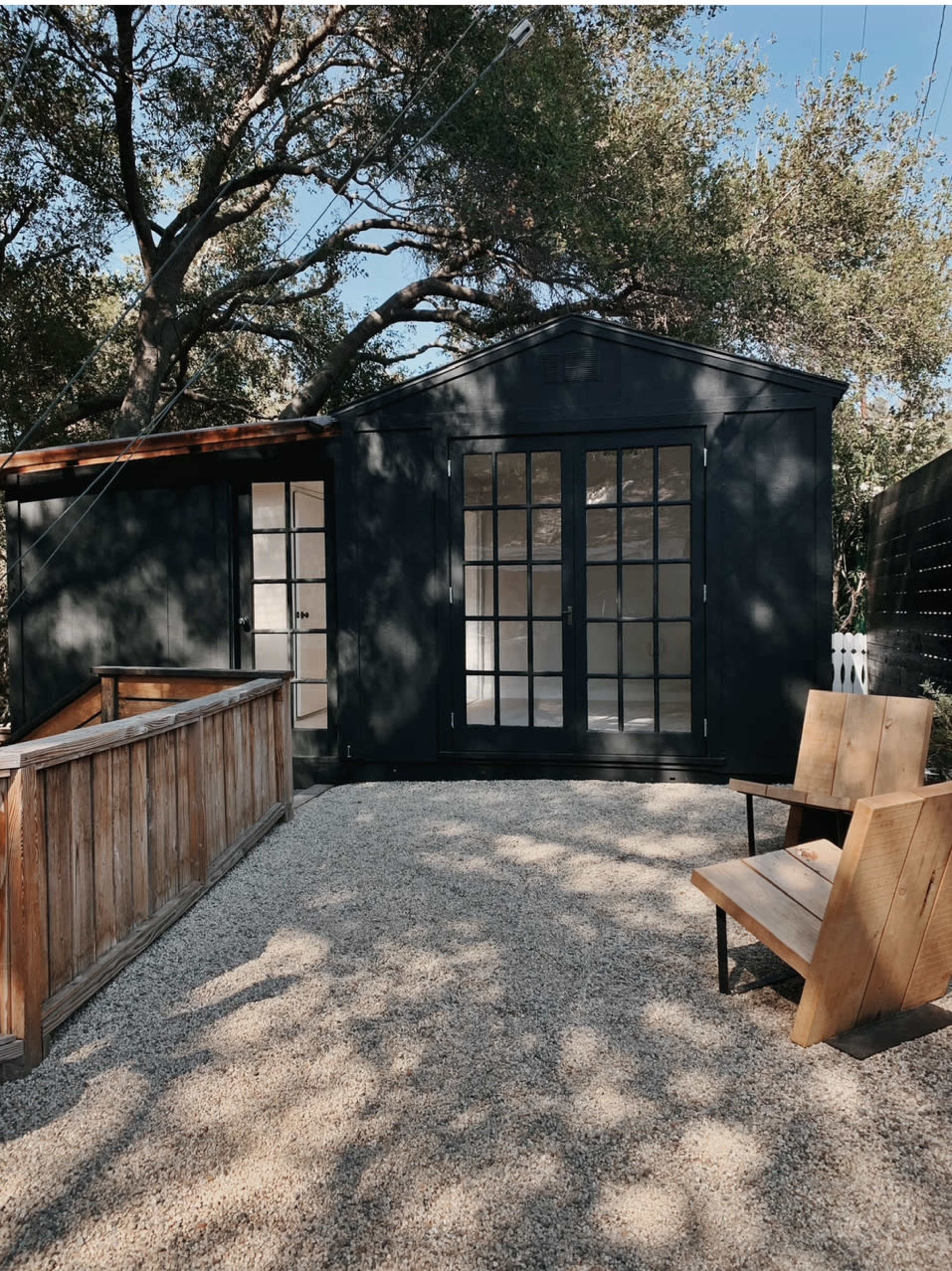 A black, modern building with large windows sits in a gravel area, accompanied by two wooden benches and surrounded by trees.