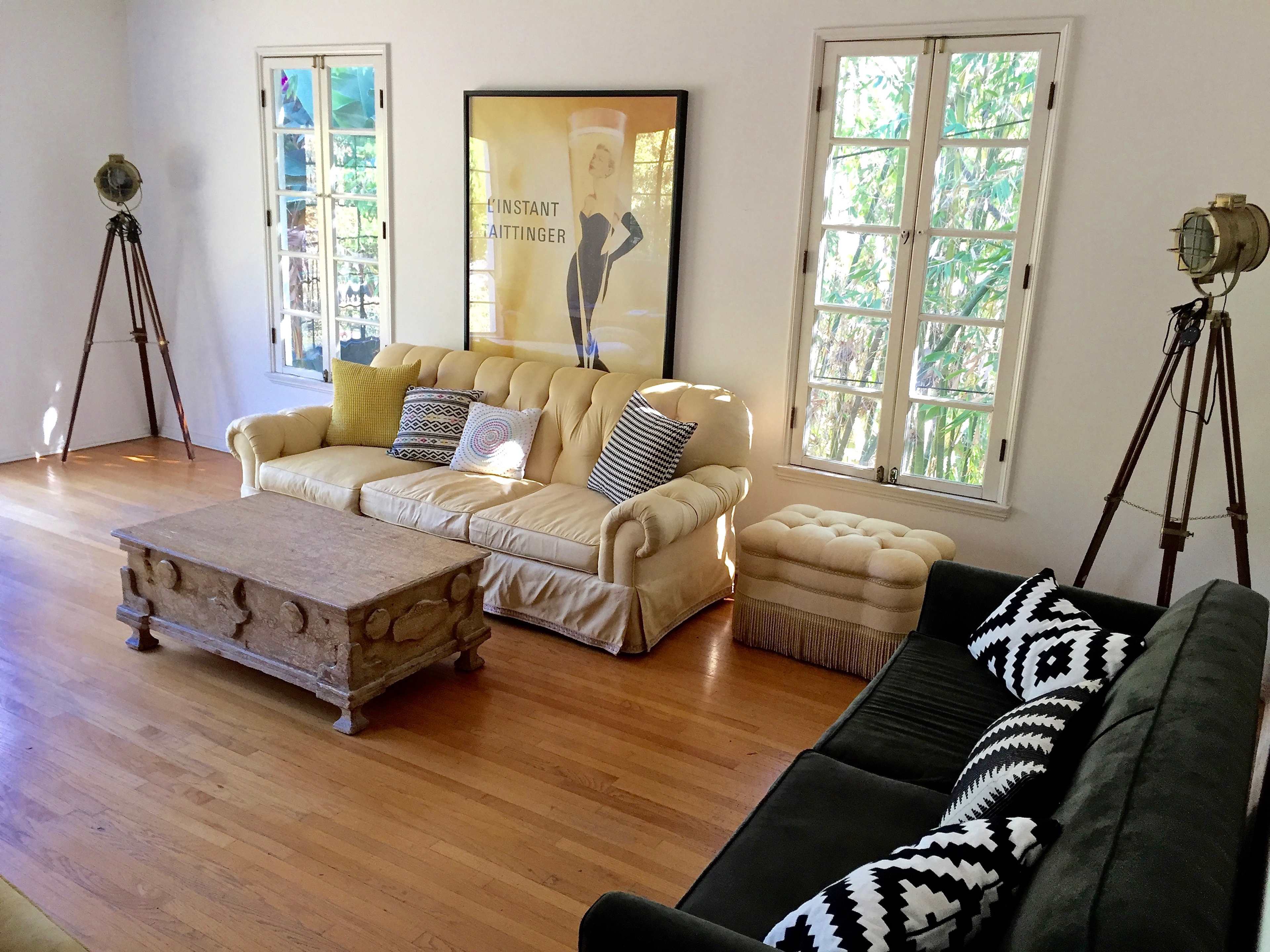 The room features two sofas, one cream-colored and one black, flanking a wooden coffee table, with vintage-style lighting and large windows allowing natural light in.