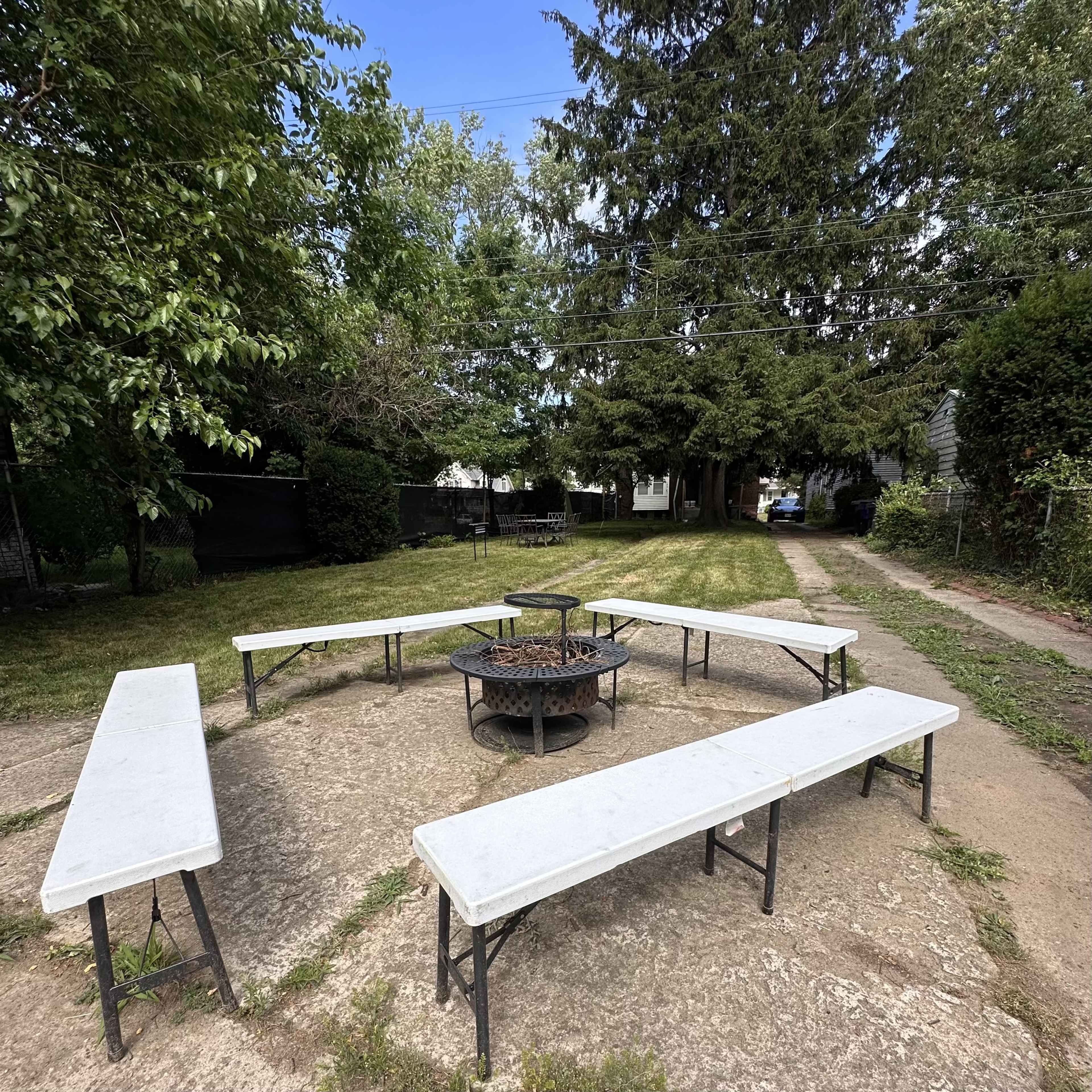 The image shows a circular stone fire pit surrounded by six white benches in a grassy yard.
