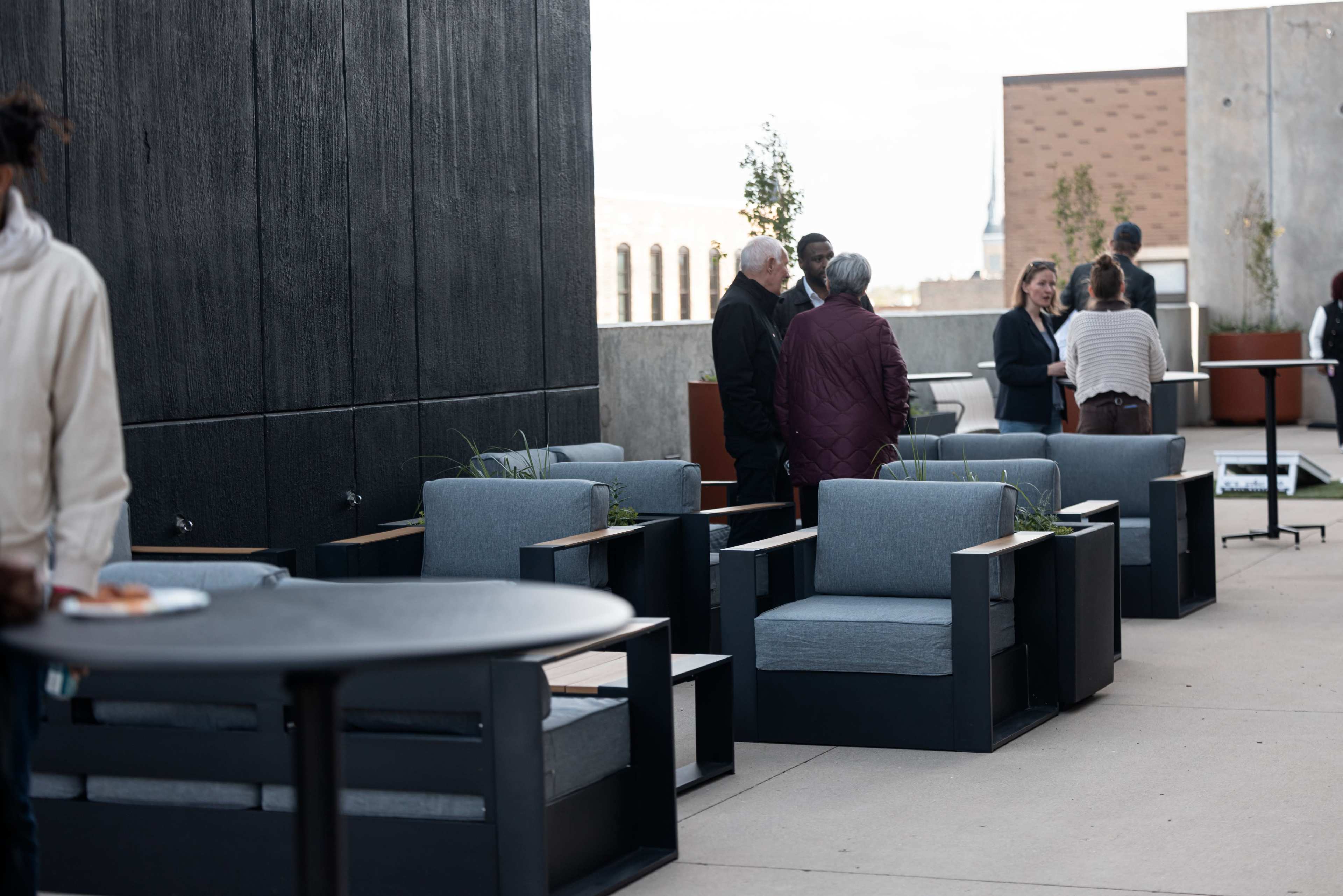 A rooftop gathering features people conversing near modern outdoor furniture, including blue chairs and small tables.
