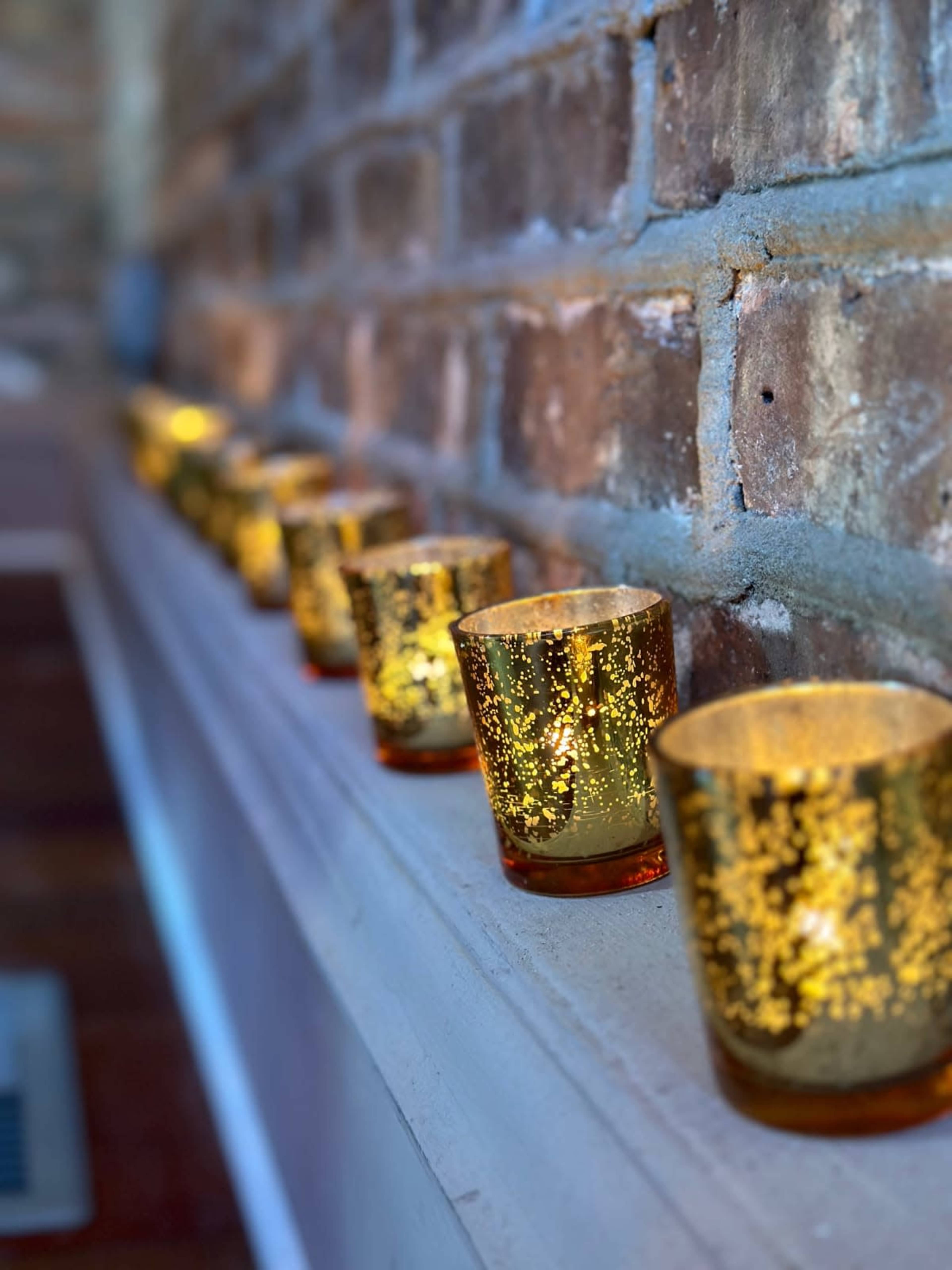 A row of golden glass candle holders with a glittery finish is arranged along a ledge in front of a brick wall.