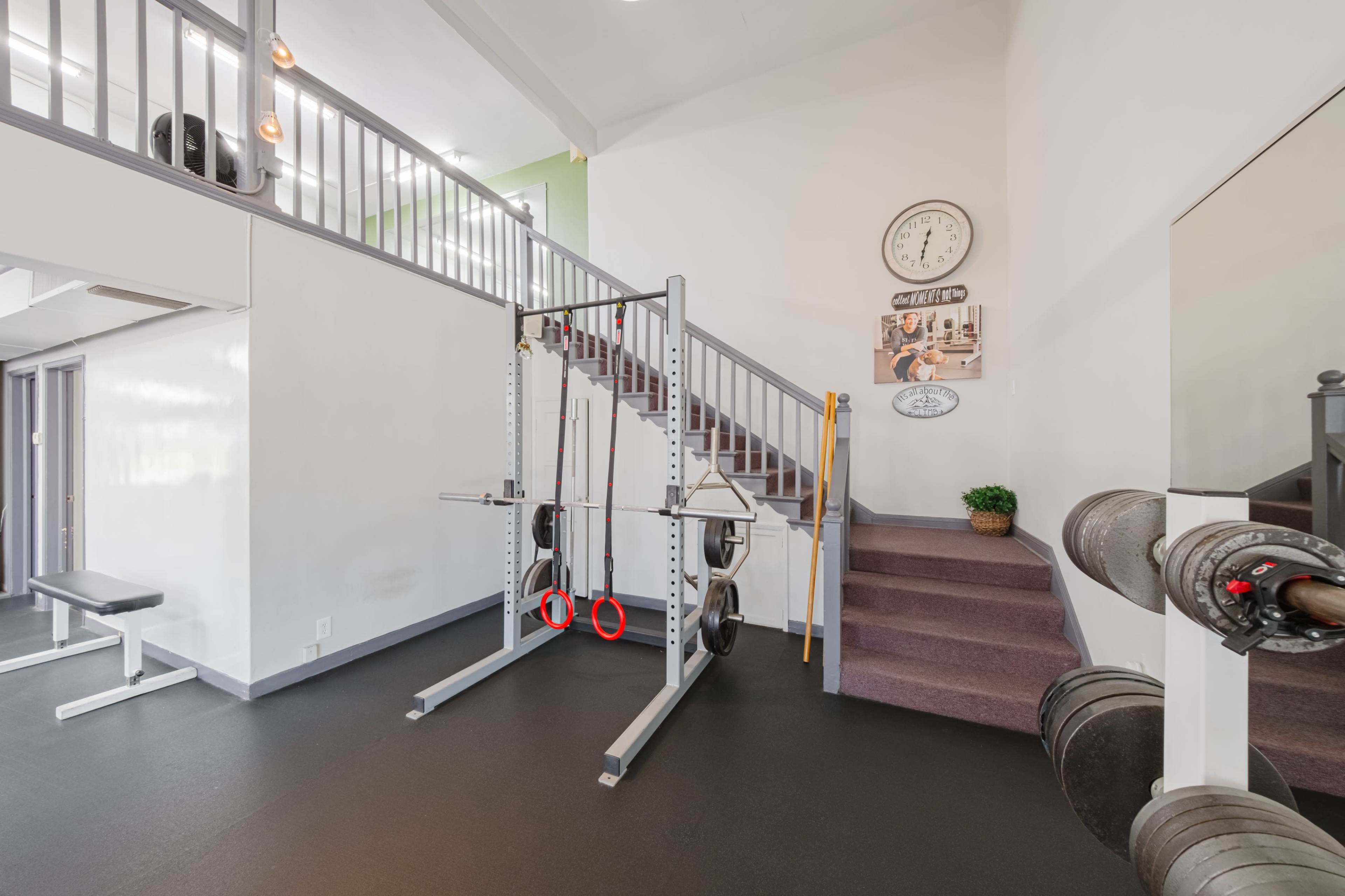 A gym area featuring a weightlifting setup with a squat rack, rings, a bench, and various weight plates positioned against the wall, alongside a staircase leading to an upper level.