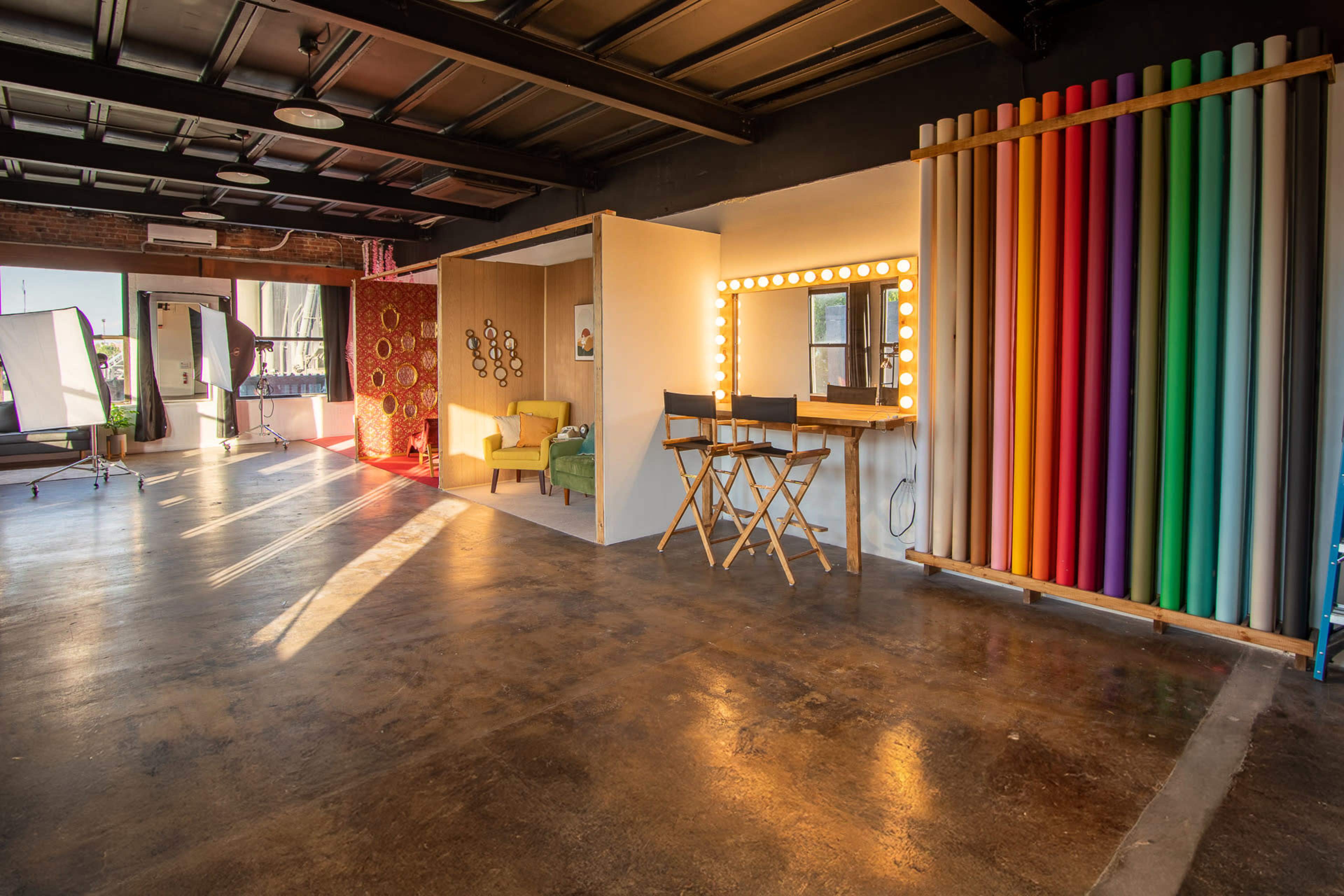 The image shows a well-lit studio space featuring a seating area, a makeup station with lights, and colorful backdrop rolls.