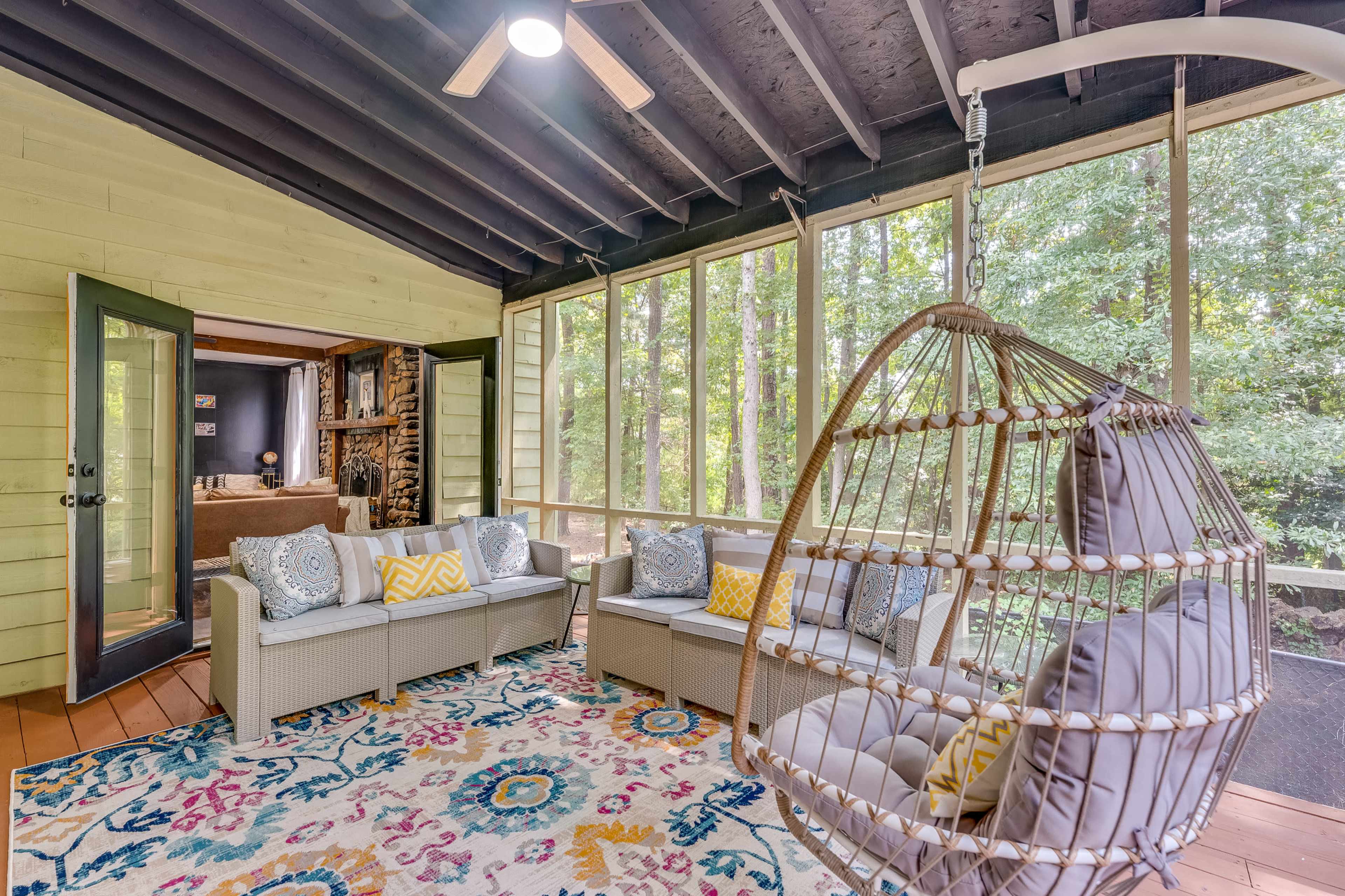 The image shows a screened-in porch with a hanging chair and woven furniture arranged on a colorful rug, surrounded by large windows that offer a view of the outdoors.