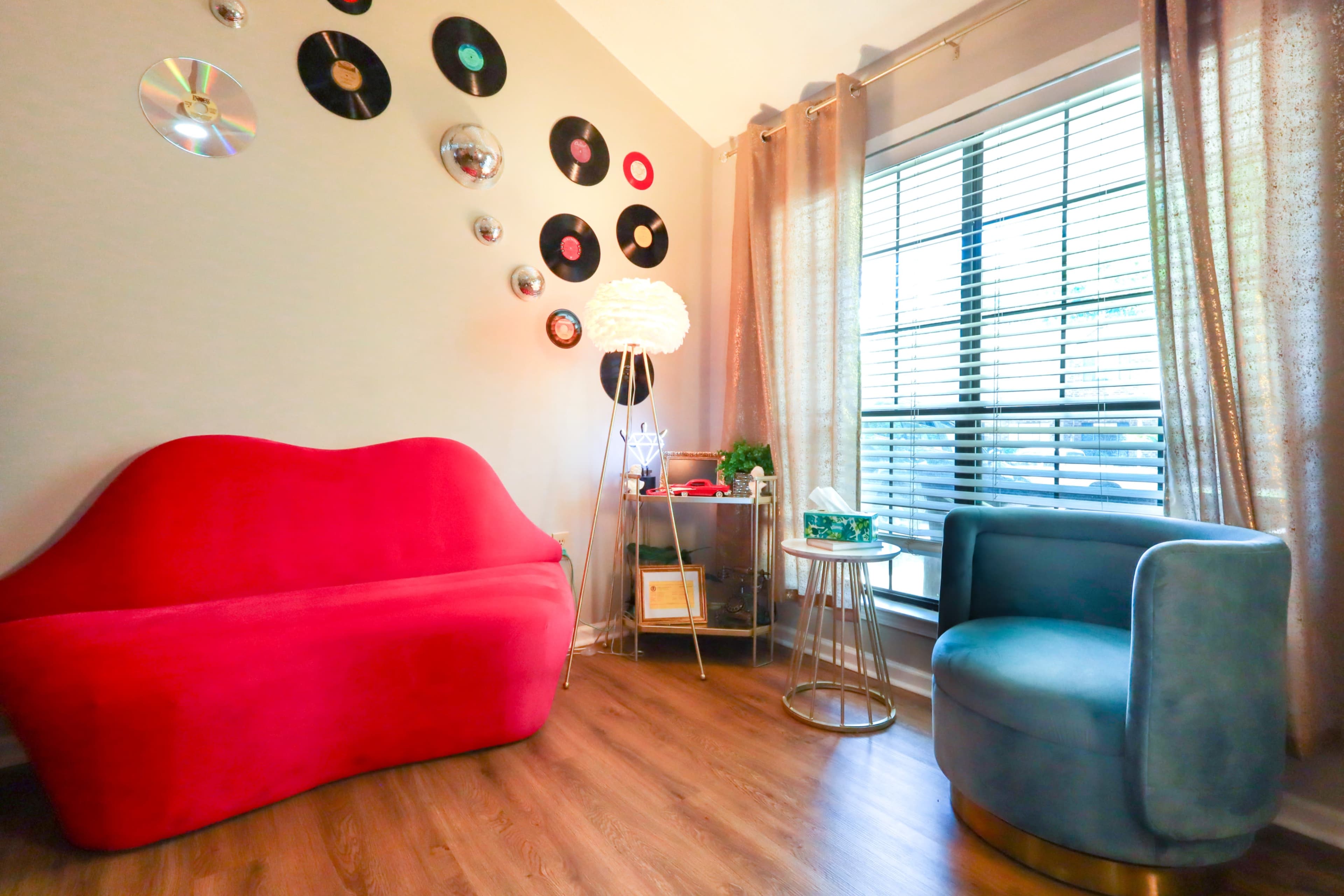 The image shows a cozy room with a red sofa and a blue chair, decorated with vinyl records on the wall and natural light streaming through a window with blinds.