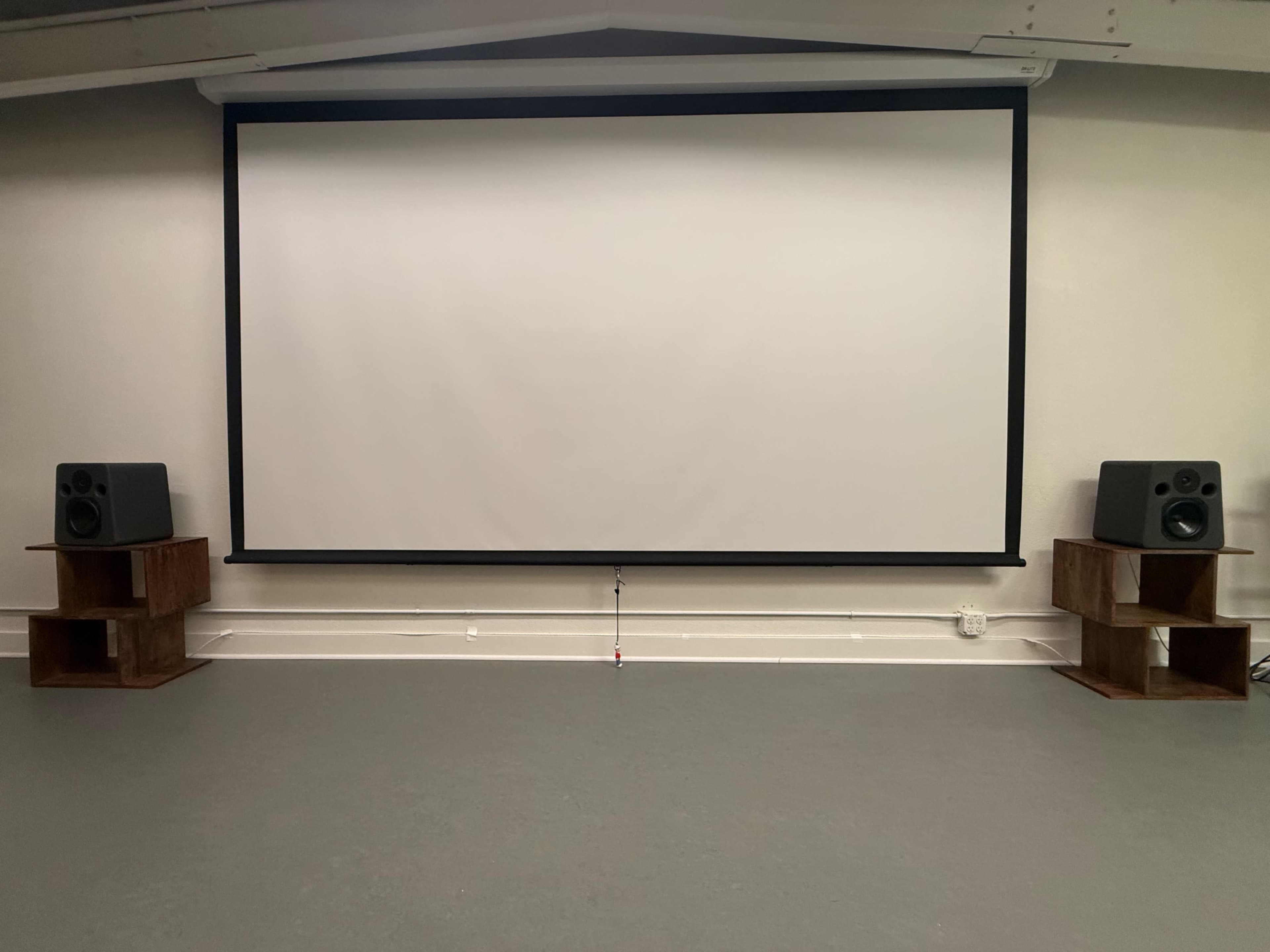 The image shows a large blank projection screen mounted on a wall, flanked by two speakers on wooden stands in a simple, open room.
