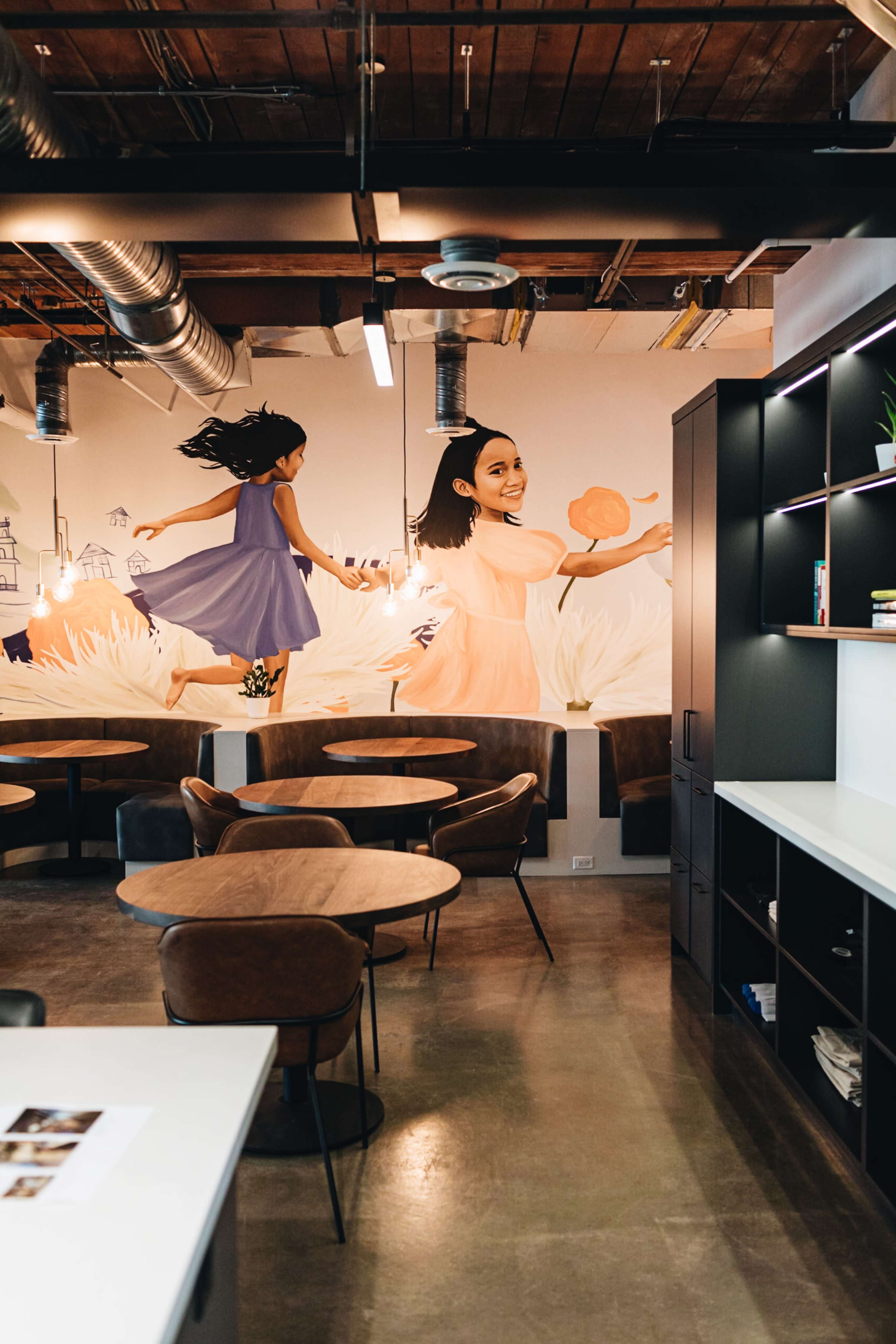 The image shows a modern workspace with wooden tables and chairs, featuring a colorful mural of two children playing on the wall.