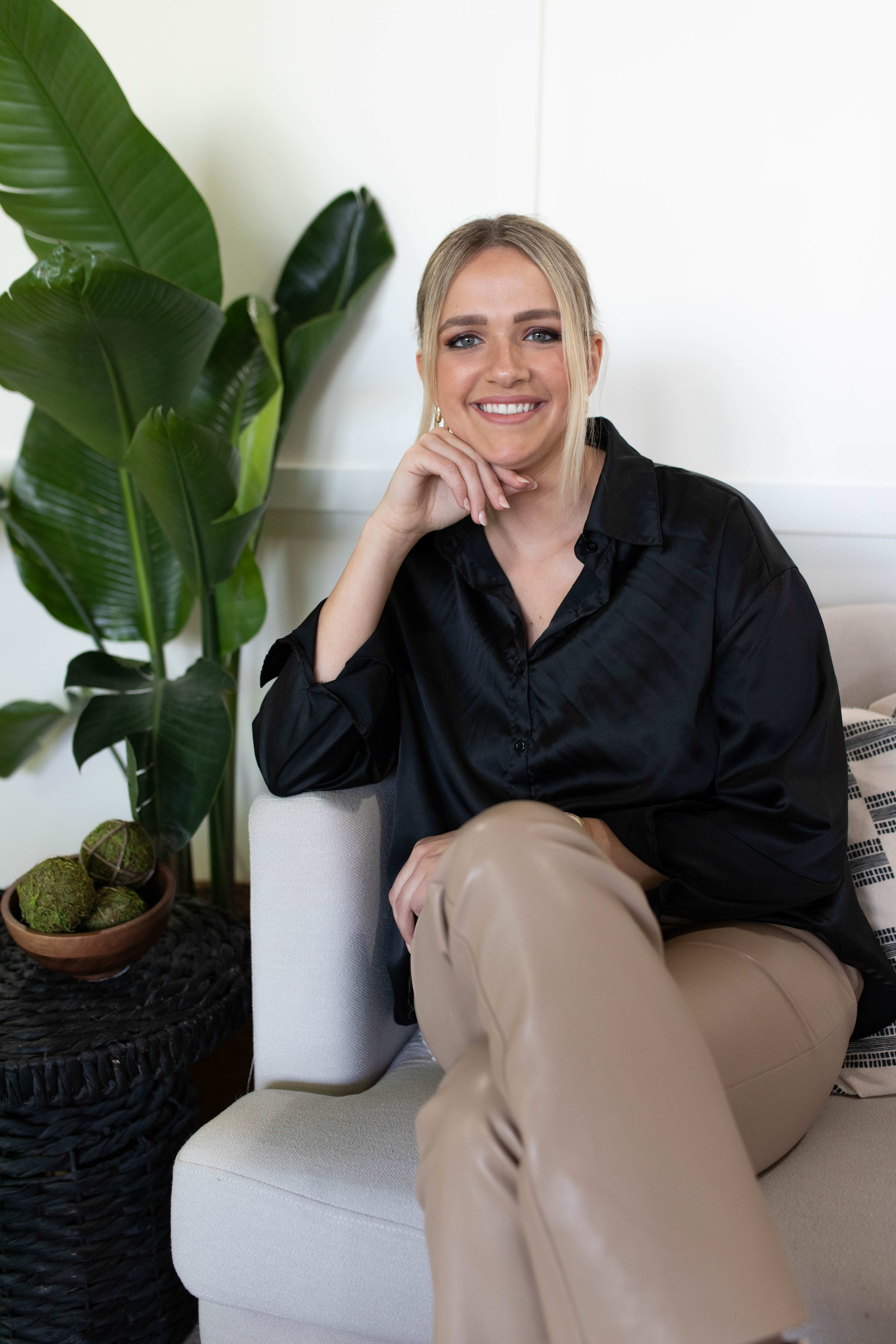 Una mujer posando para un retrato en un sofá beige junto a una planta verde en una maceta.