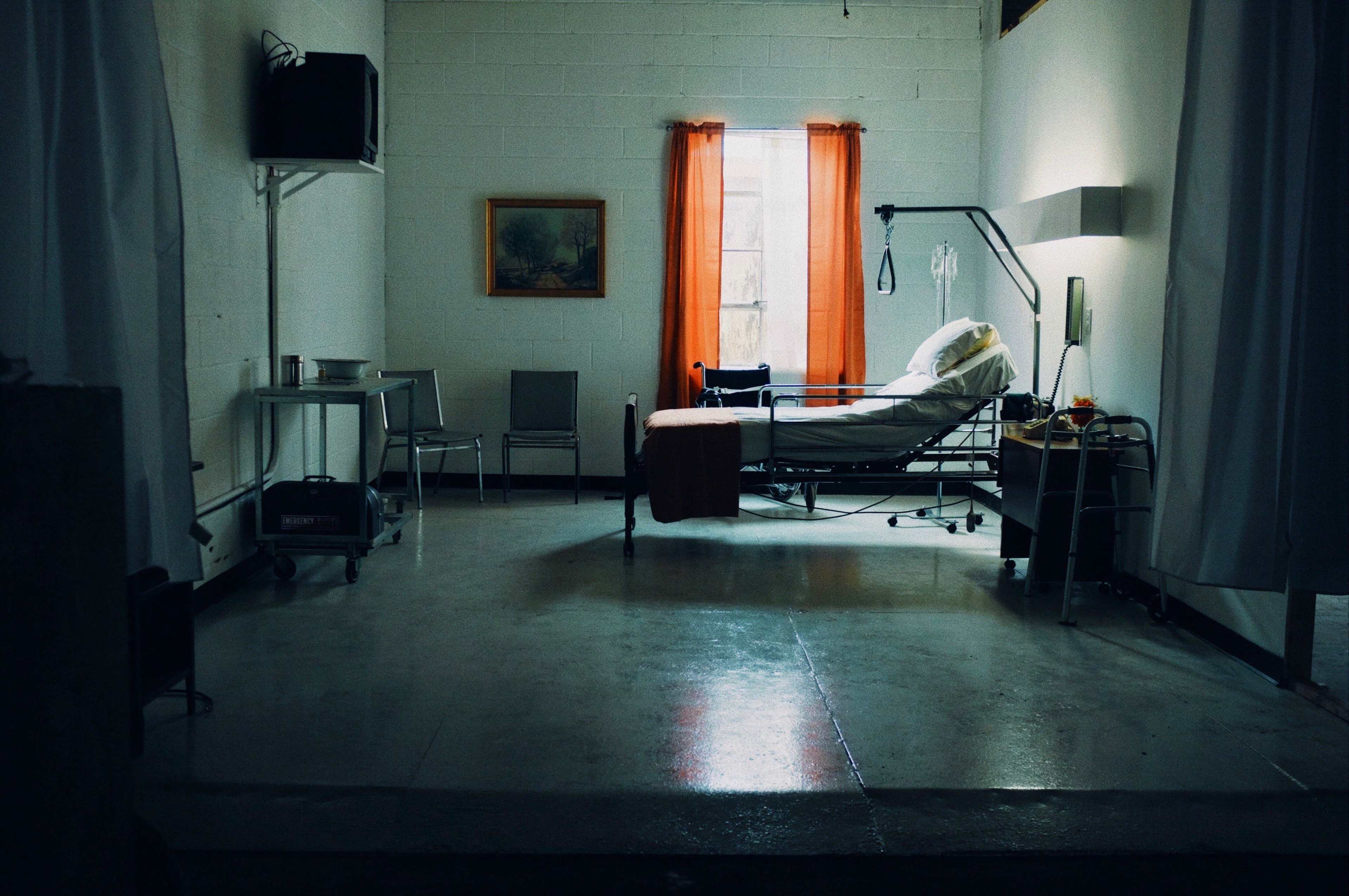 The image shows a sparse hospital room with a bed, medical equipment, and orange curtains by a window.