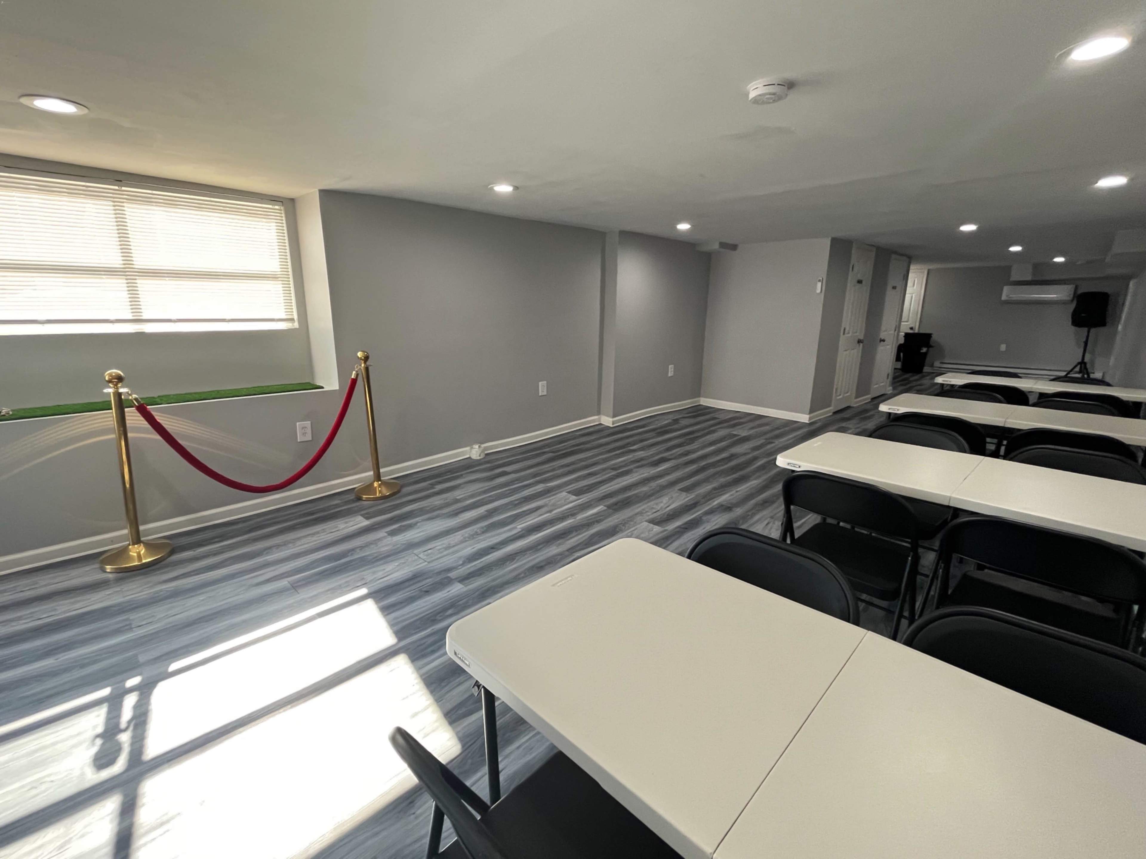 A well-lit, empty room with gray walls, chairs, and tables arranged for an event, along with a red rope barrier near a window.