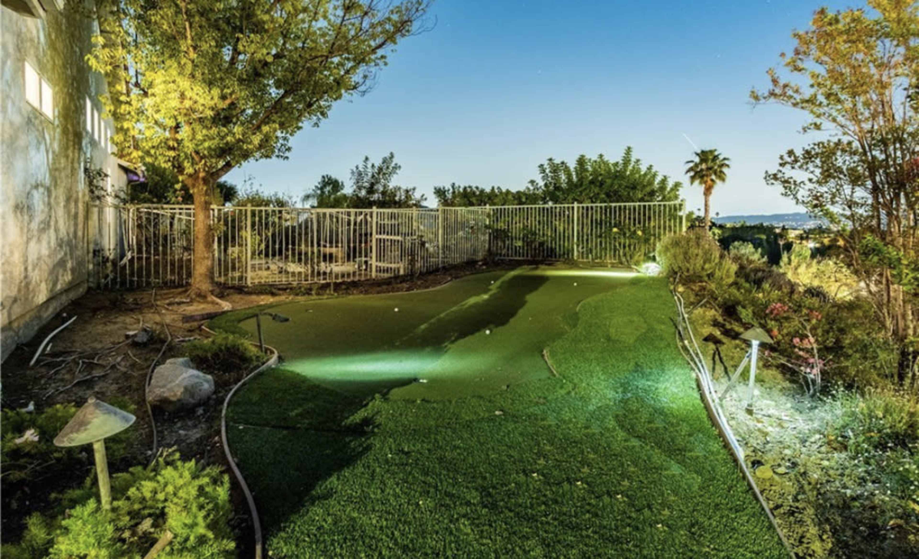A fenced backyard features a small putting green illuminated by lights, surrounded by trees and shrubs.