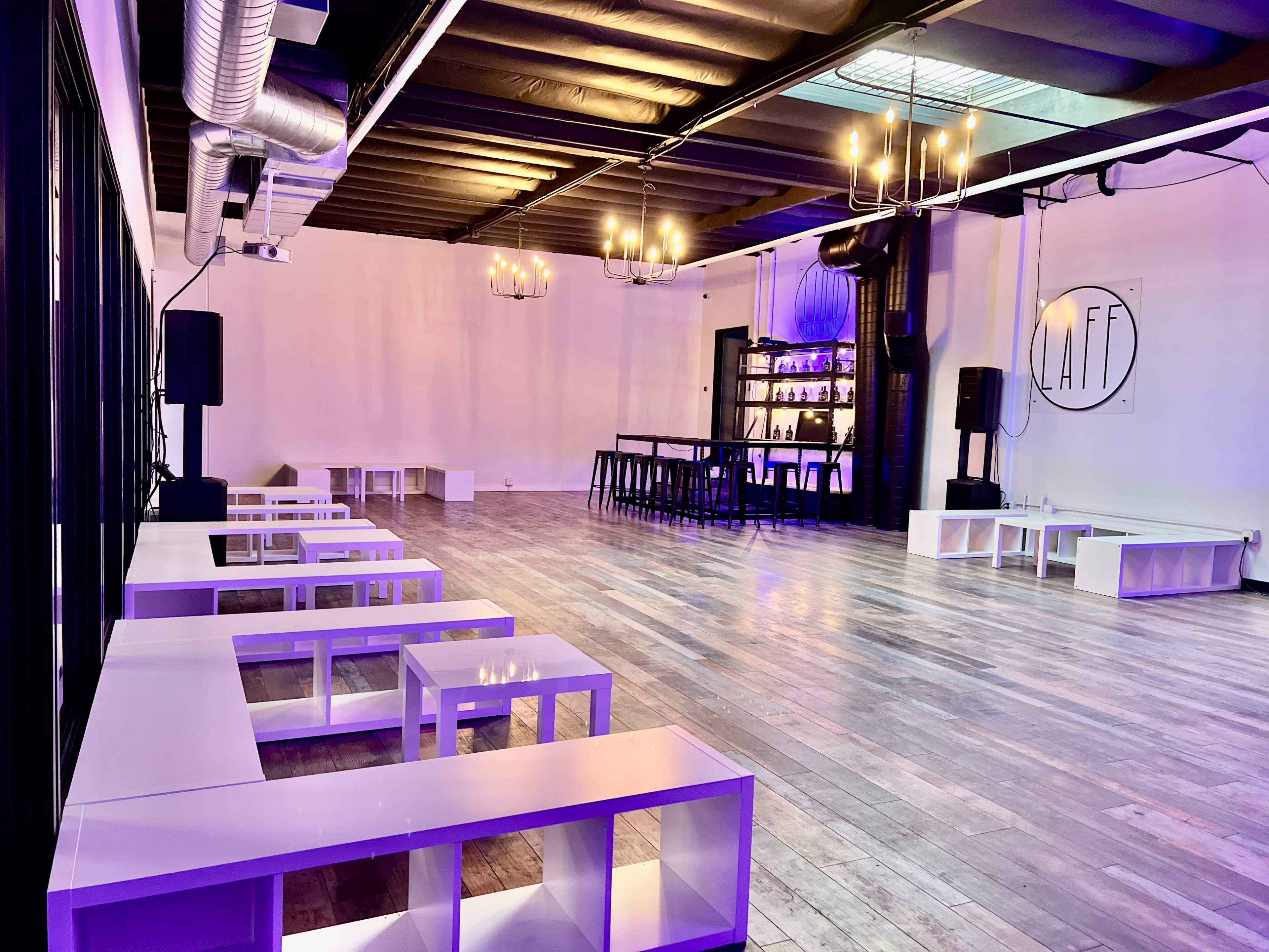 A spacious event venue with wooden flooring, white low tables, and a bar area illuminated by purple lighting and chandeliers.