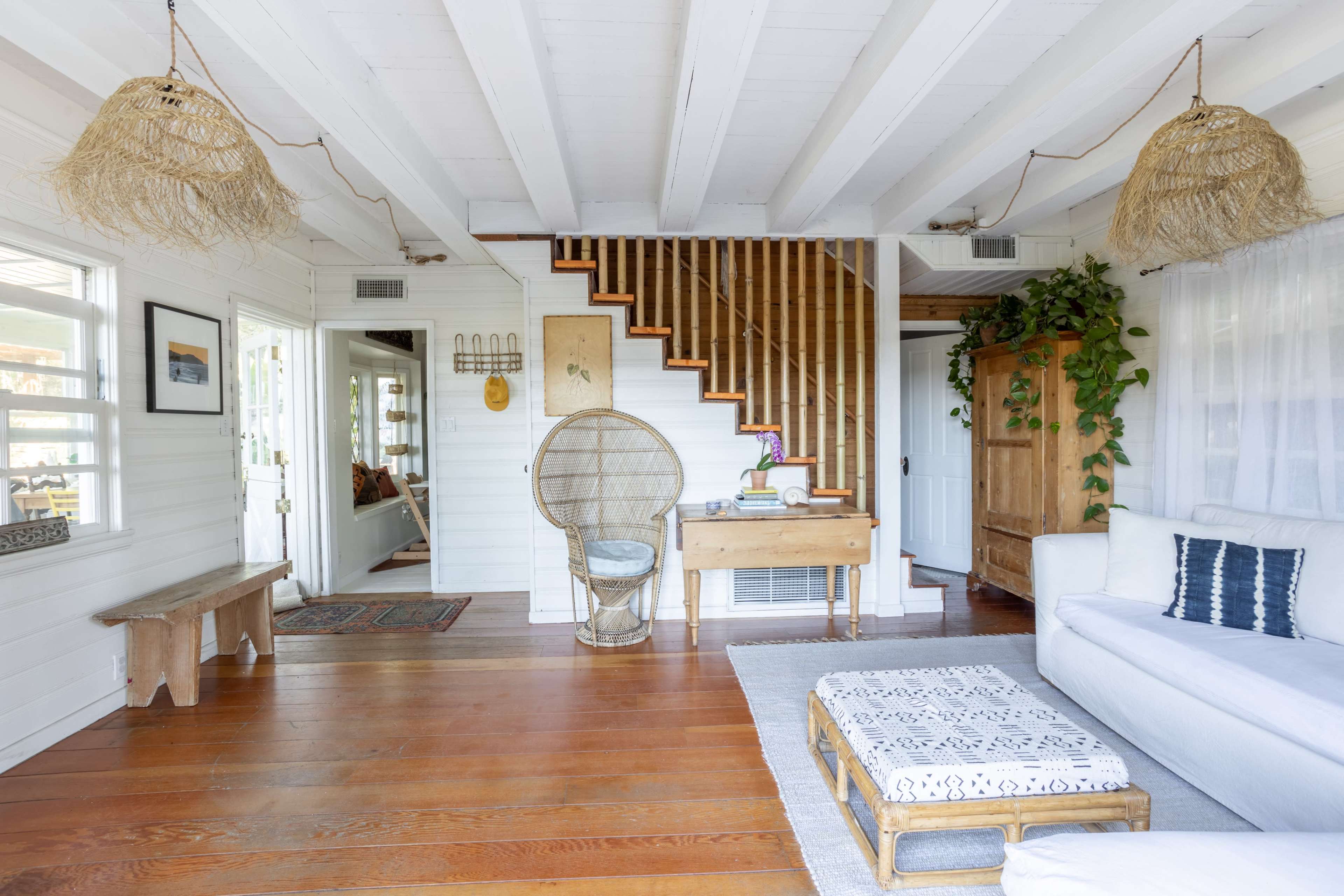 A cozy, well-lit living space with white walls, wooden floors, a staircase, and a mix of natural elements such as woven light fixtures and plants.