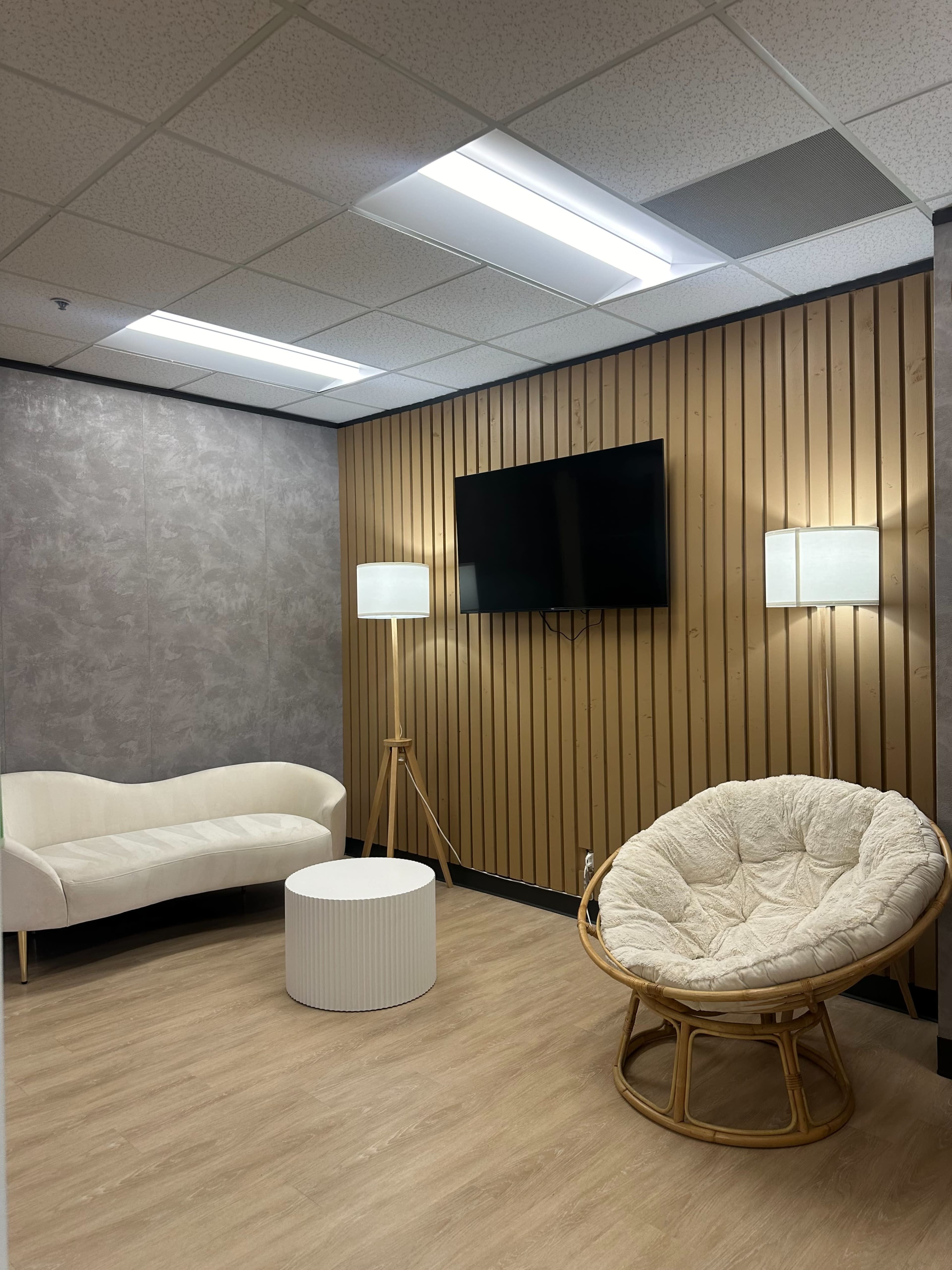 A small indoor lounge area with a white couch, a round coffee table, a tall floor lamp, a wall-mounted television, and a round, cushioned chair.